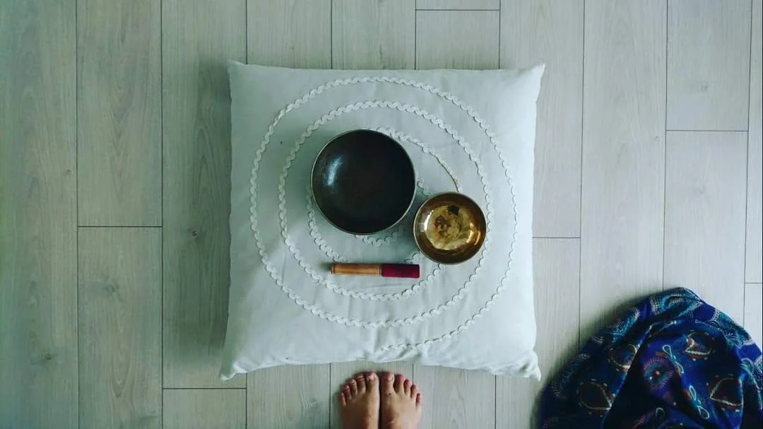 A meditation setup on a white cushion with concentric circles in a spiral pattern, featuring a black meditation bowl, a smaller brass bowl, a red lighter, and a stick of incense. A person's bare feet are visible at the bottom of the image on a light 
