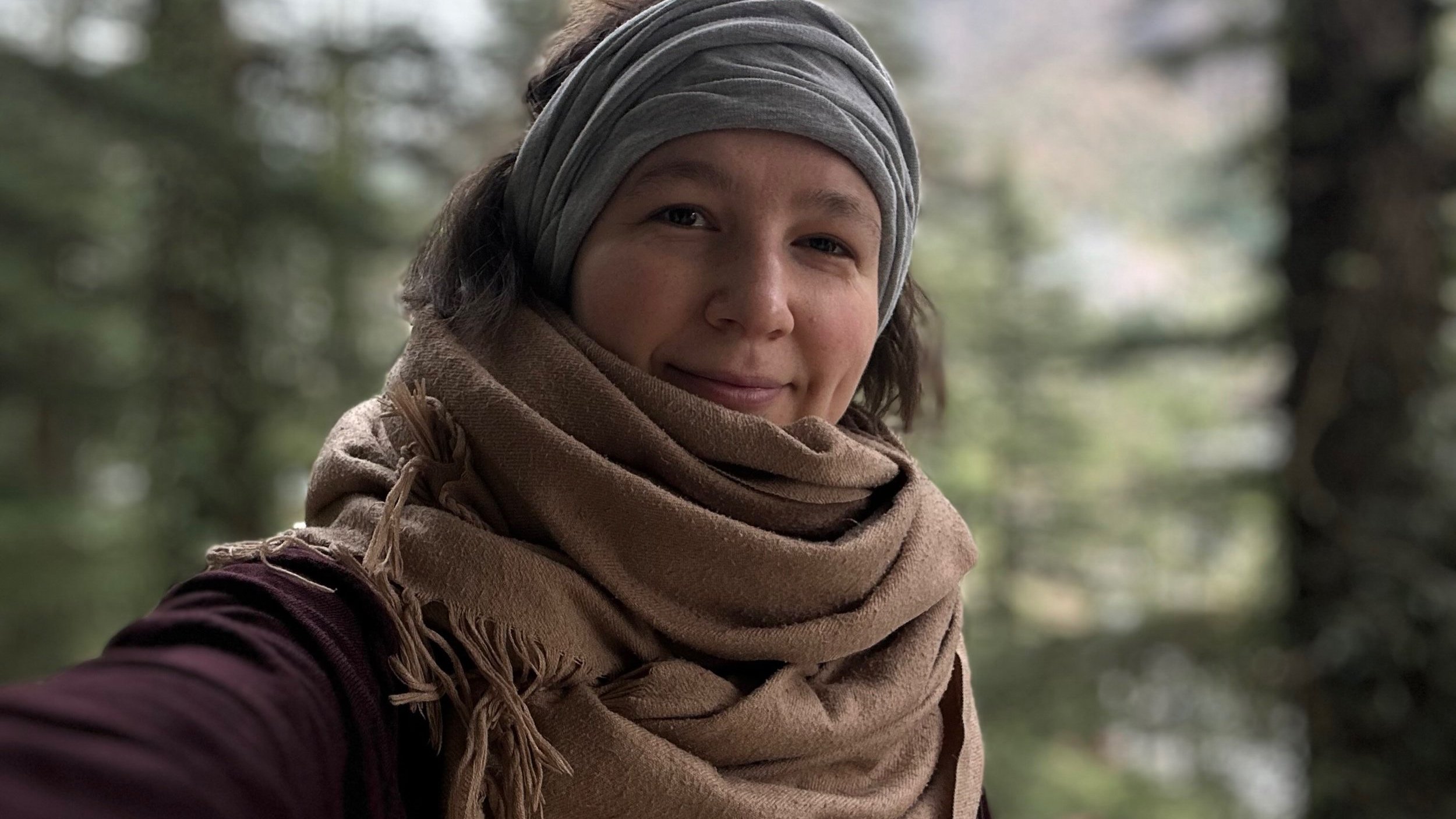 A person wearing a gray headwrap and tan scarf, looking at the camera with a slight smile, in a forested area.