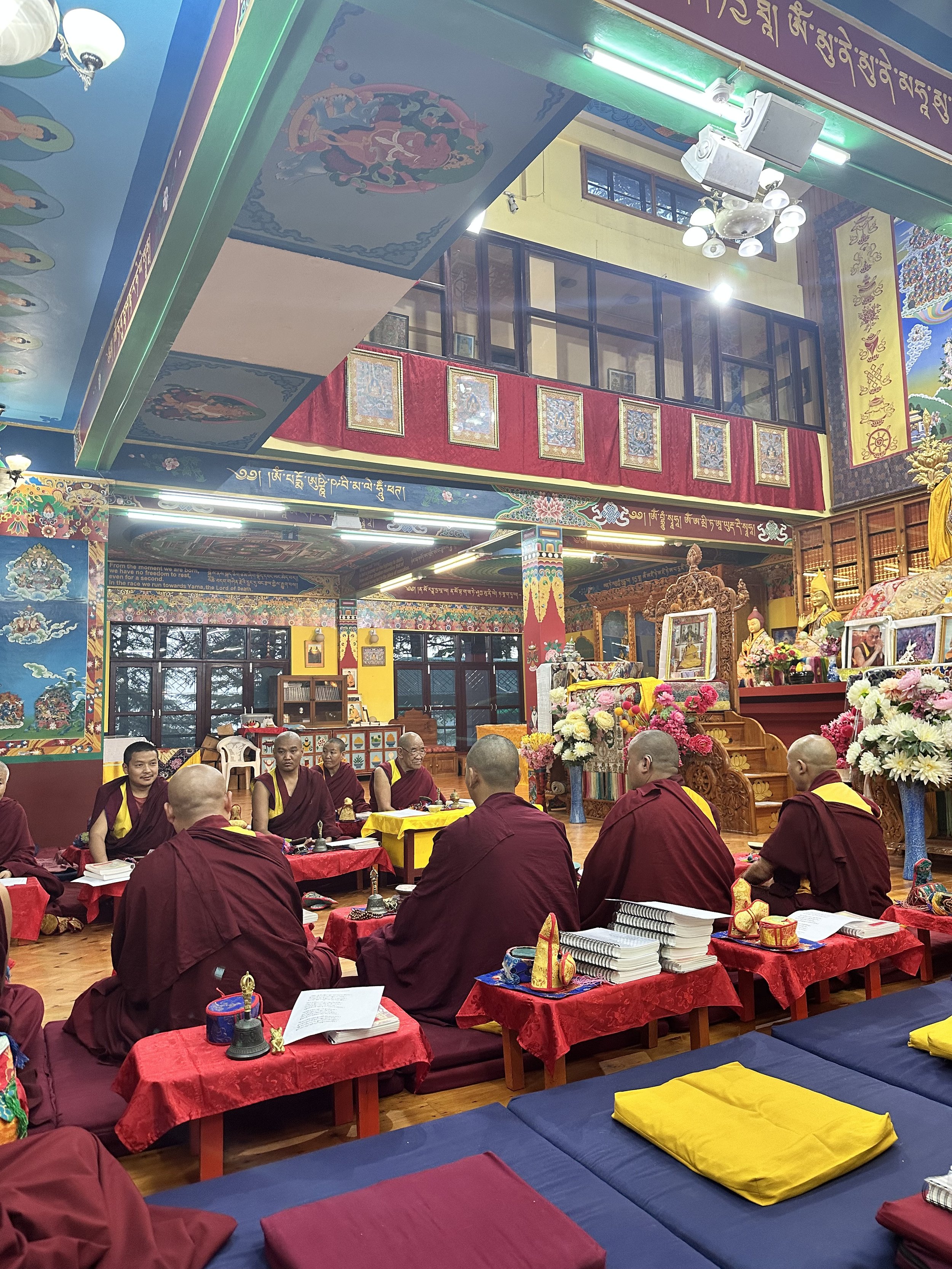 Monks in maroon robes sitting in a brightly decorated Buddhist temple, engaged in a ceremony or study session with religious texts and items on tables.