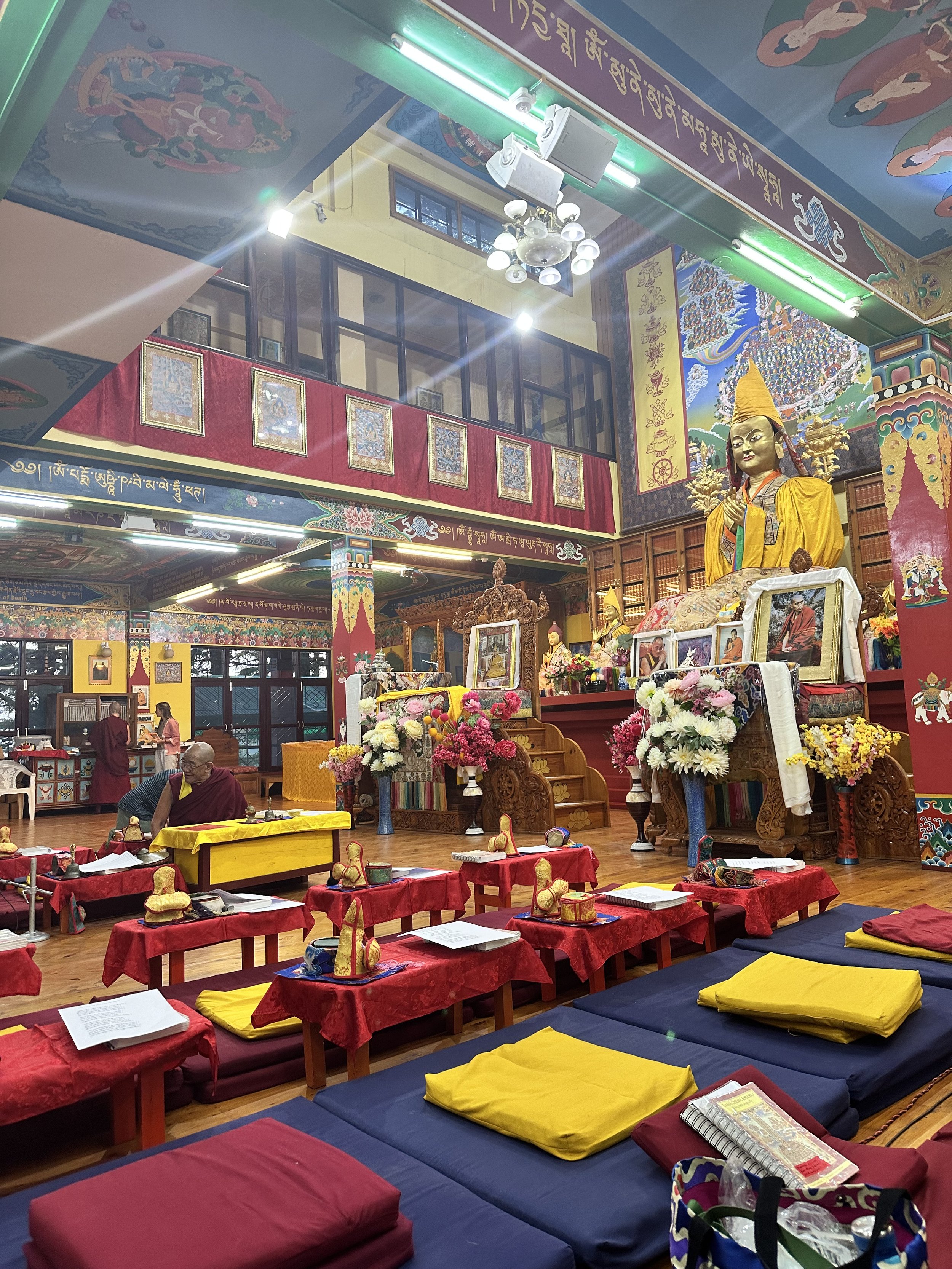Interior of a colorful Buddhist temple with a large statue, decorated altar, cushions, and prayer tables. Monks are present, and the walls are adorned with traditional artwork and scripture.