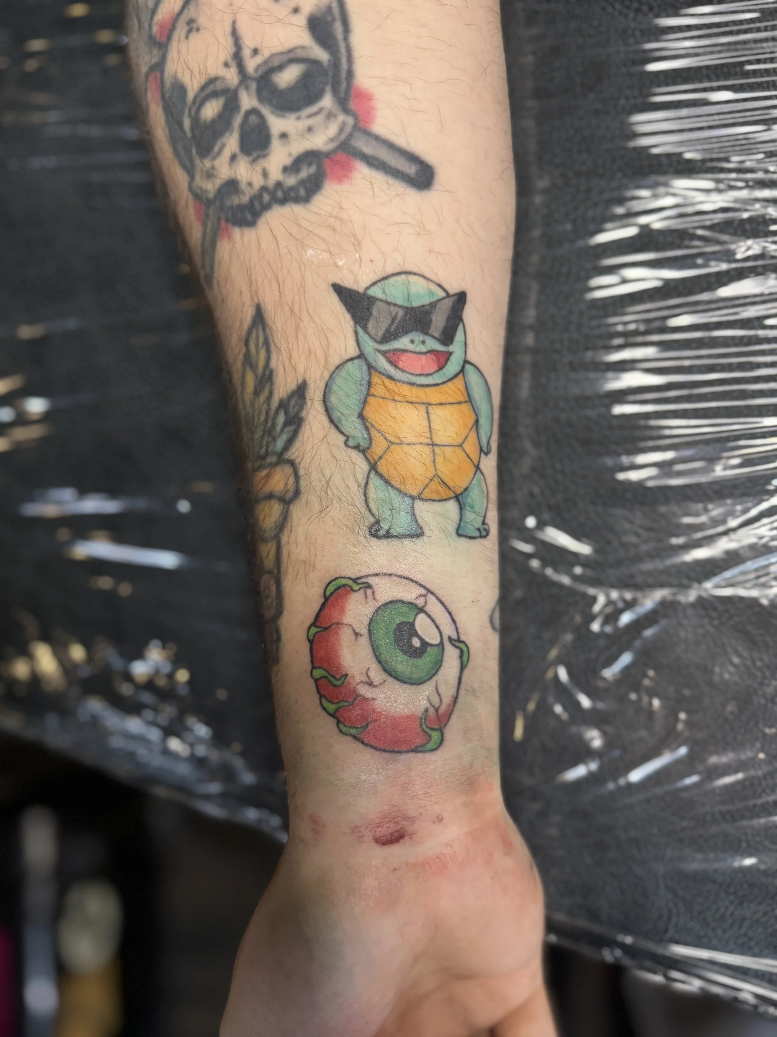 Tattoo of a cartoon turtle wearing sunglasses, with a shell and a wide smile, alongside a cartoon eye with a red, fiery appearance.