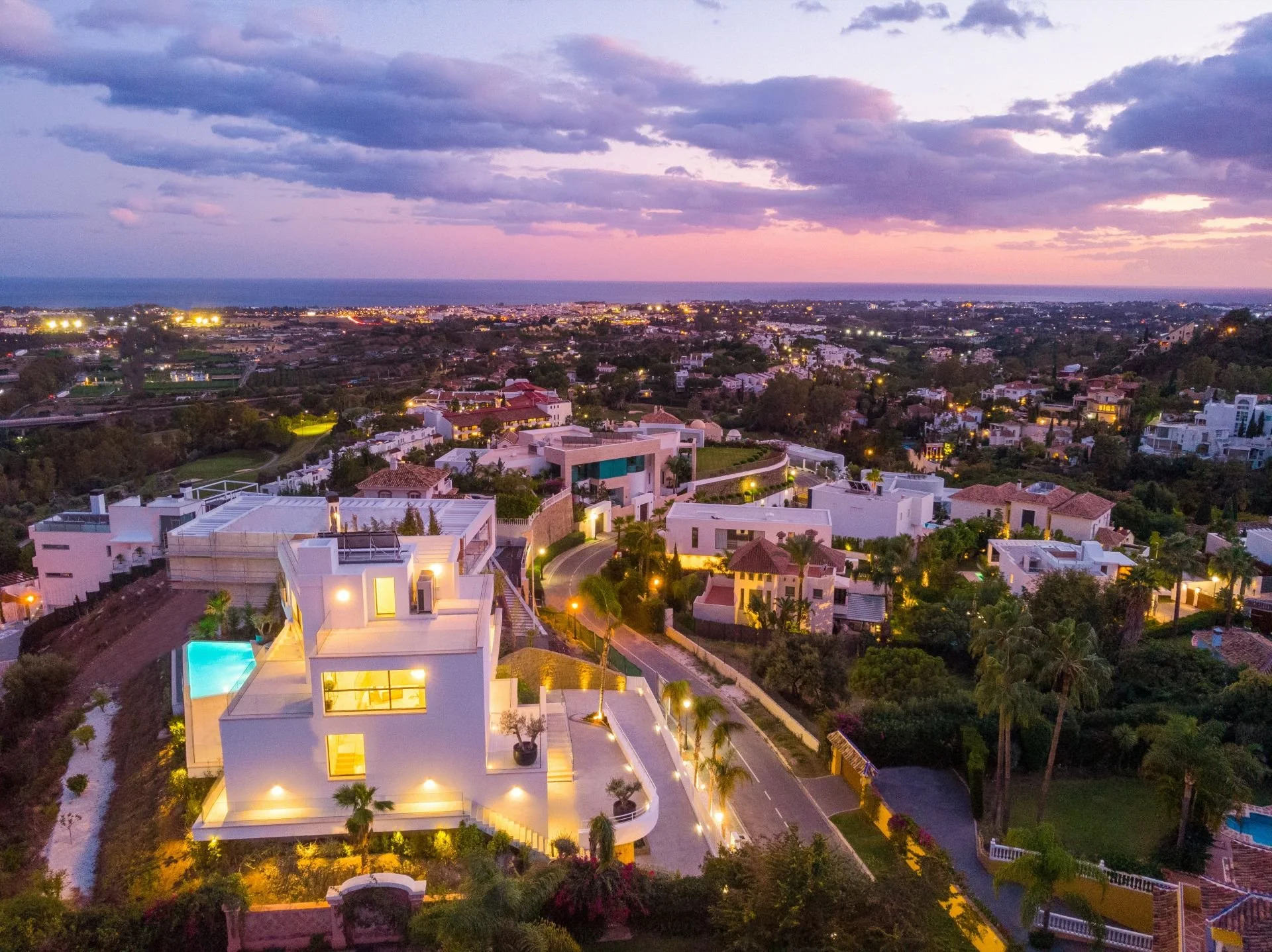 An aerial view of a coastal city at dusk showing illuminated modern houses, some with pools, greenery, and winding roads with streetlights, with the ocean and sunset sky in the background.