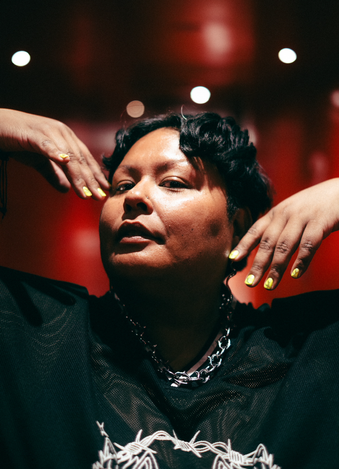 Headshot of Mar, a brown Latino person, with short wavy black hair, holding their hands by their face in a pose, wearing a silver chain and dark shirt against a red background
