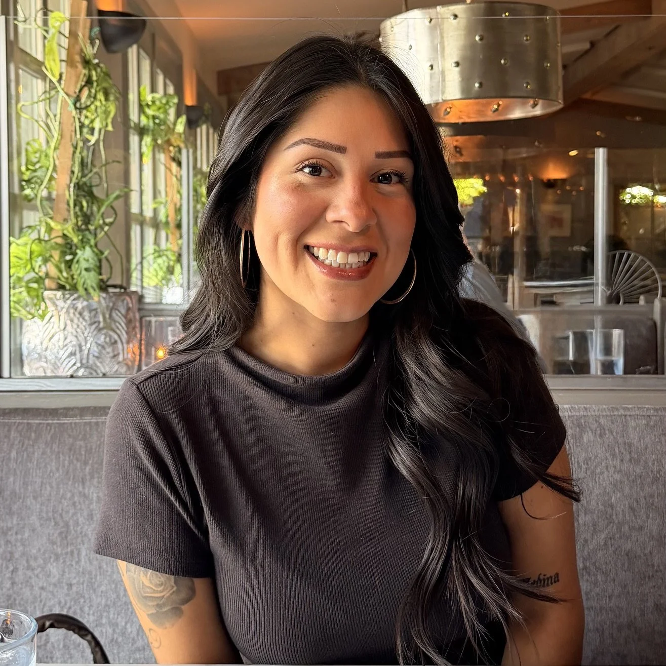 Photo of a brown Mexican woman with long wavy black hair, smiling, wearing thin hoops and a black t-shirt