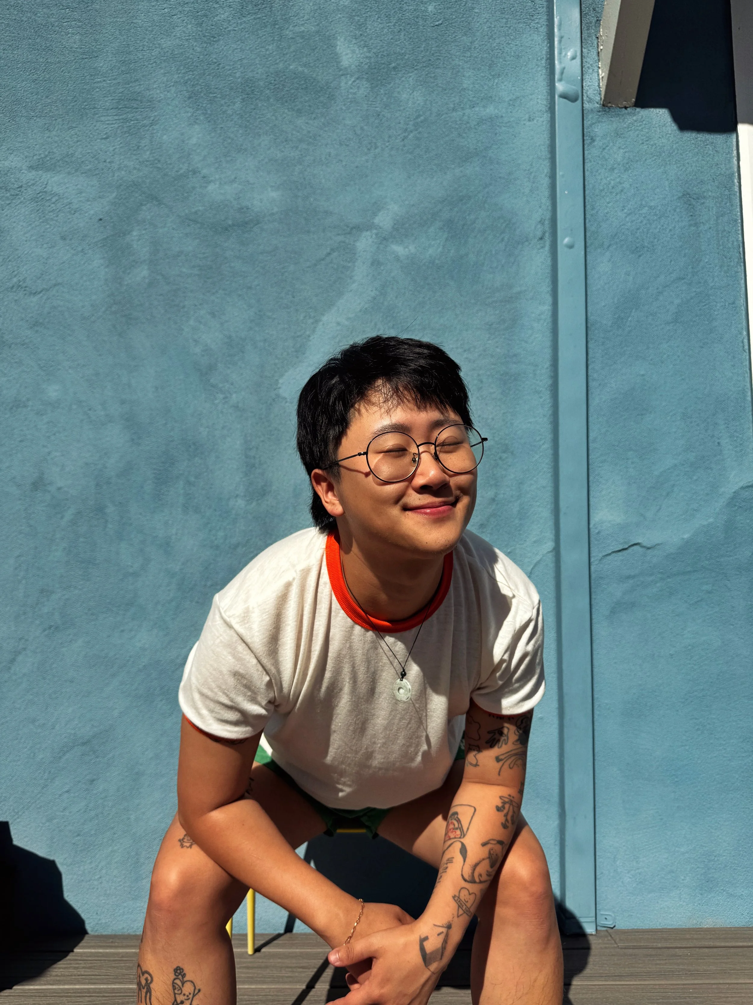 Sam is a Korean person with short black hair and black glasses. He is wearing a white t-shirt and sitting against a blue wall.