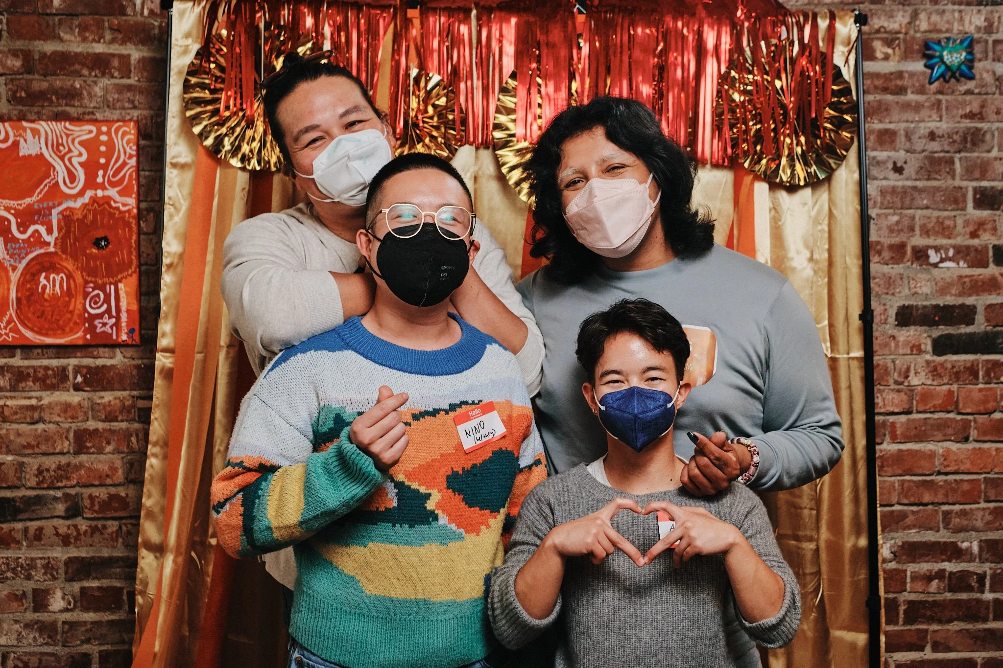 four masked people of various heights, making hearts with their hands, in front of a gold and red photobooth backdrop posing