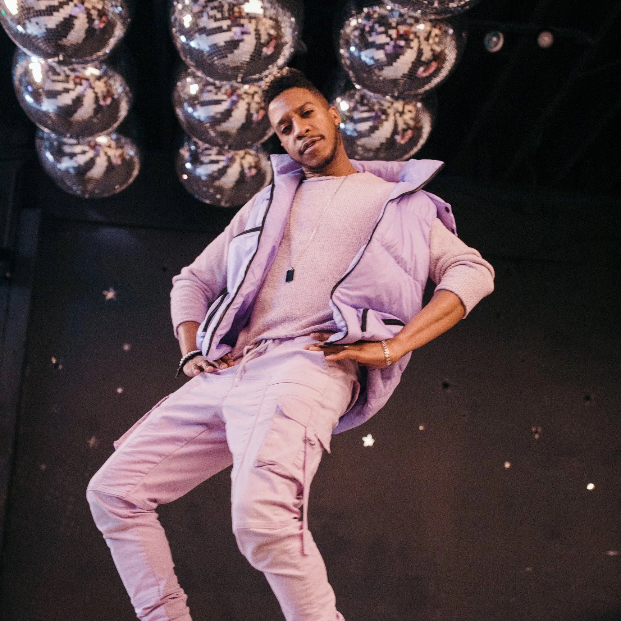 Person wearing all lilac pants, shirt and zip up vest with hands on hips, brown skin, and several disco balls in the background