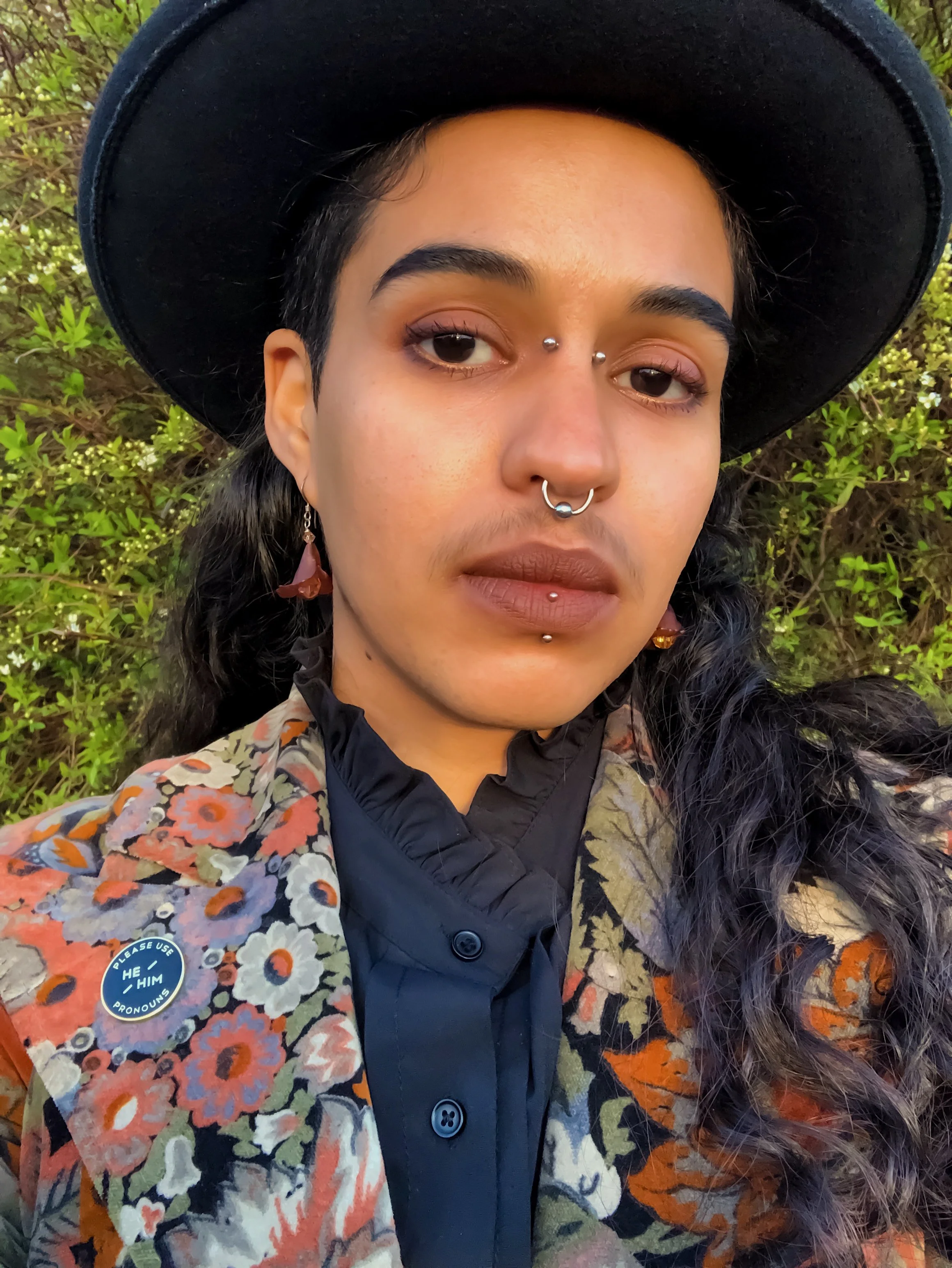 Jonathan is wearing a black hat, silver face and nose rings, and floral earrings. They are a light-skinned Black person with long dark hair and are looking into the camera