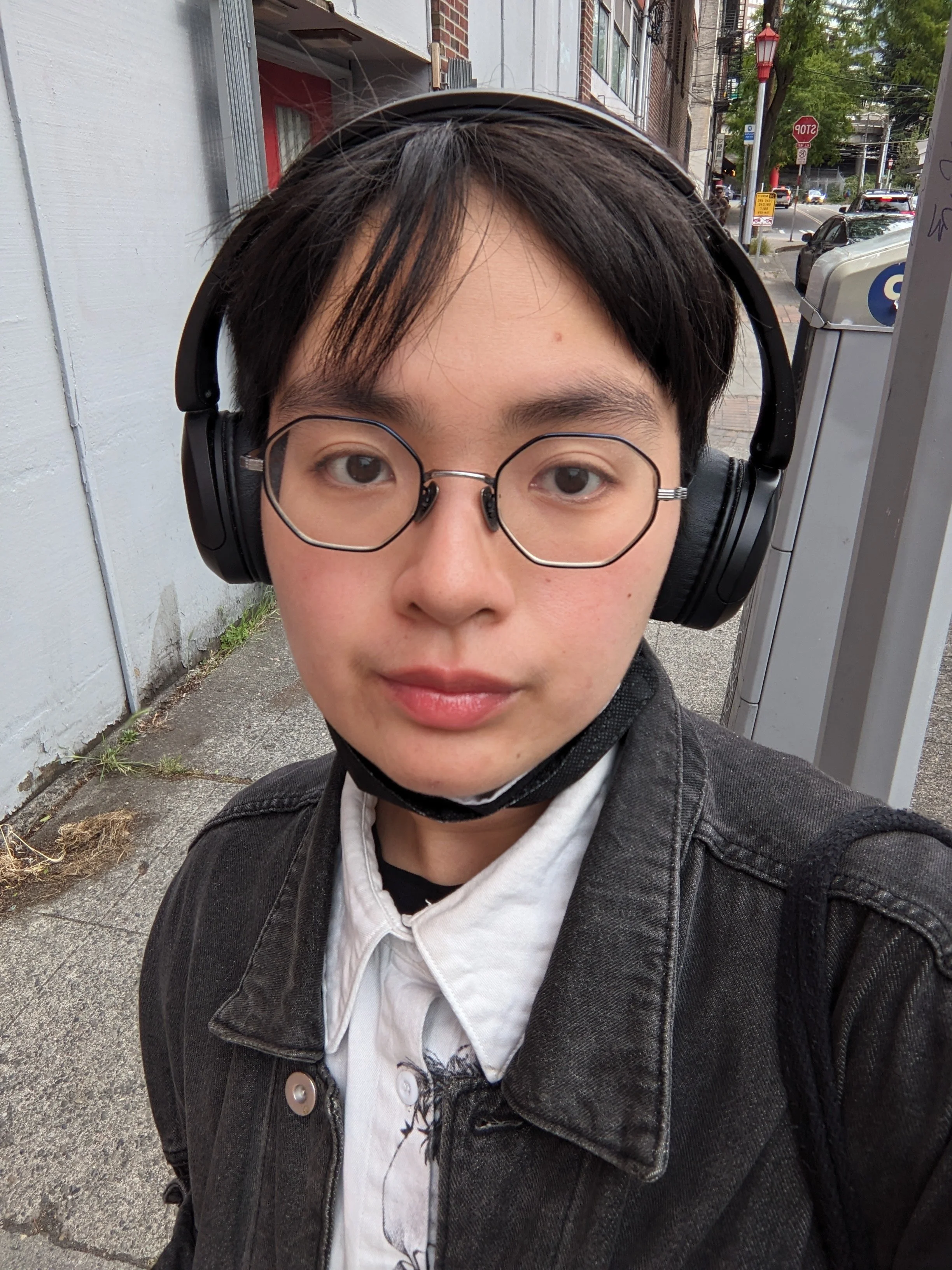 Selfie of a Vietnamese person wearing glasses, overwear headphones, a white button down, and black denim jacket. They have short black hair and dark eyes.