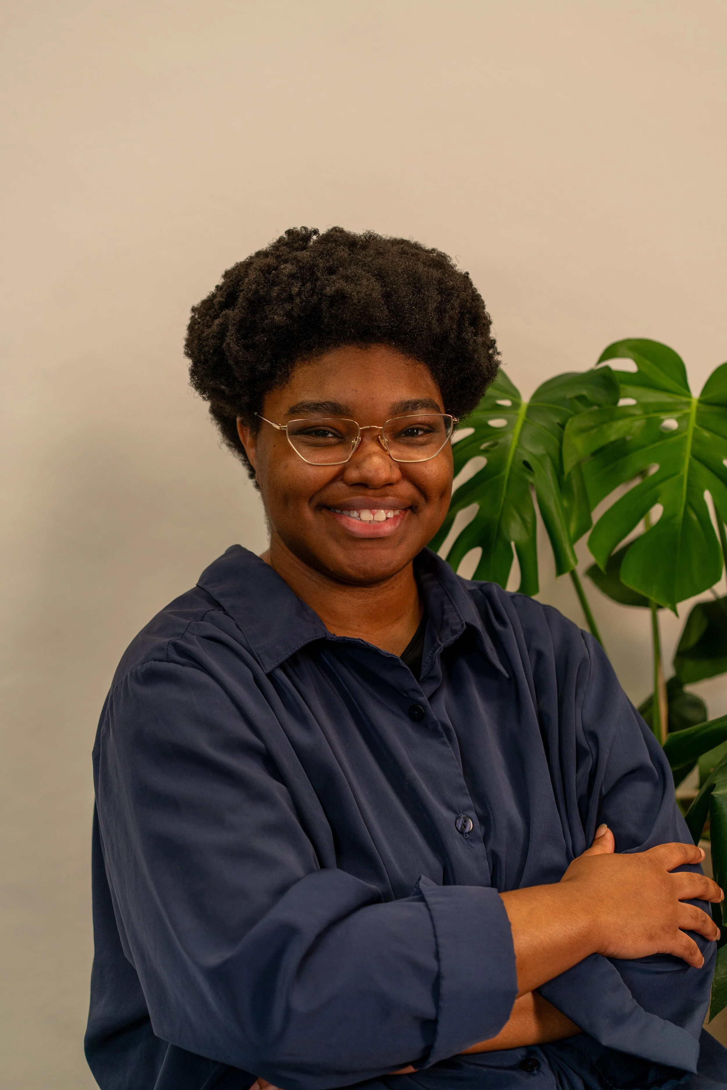 Photo of Kory, a Black person with glasses and a small afro, smiling, wearing a blue button down t-shirt, with a monstera behind them