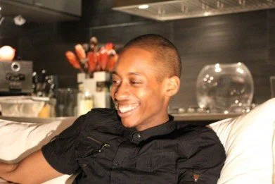 young Black man wearing Black collared shirt smiling and looking off camera to the right