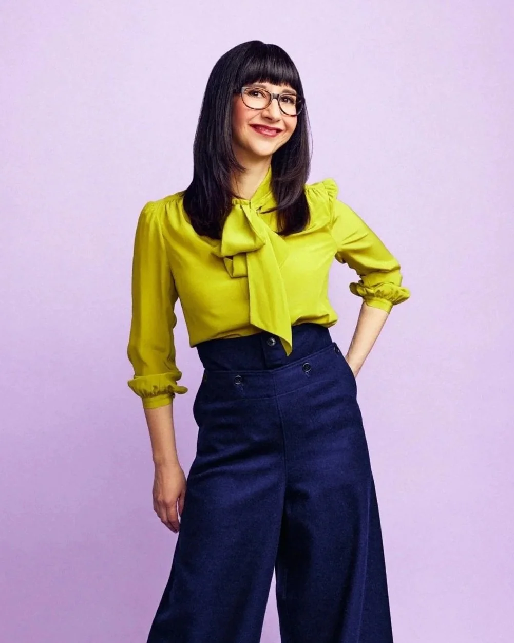 A woman with dark hair and glasses wearing a yellow blouse with a bow tie, and high-waisted dark wide-leg pants, standing against a plain purple background.
