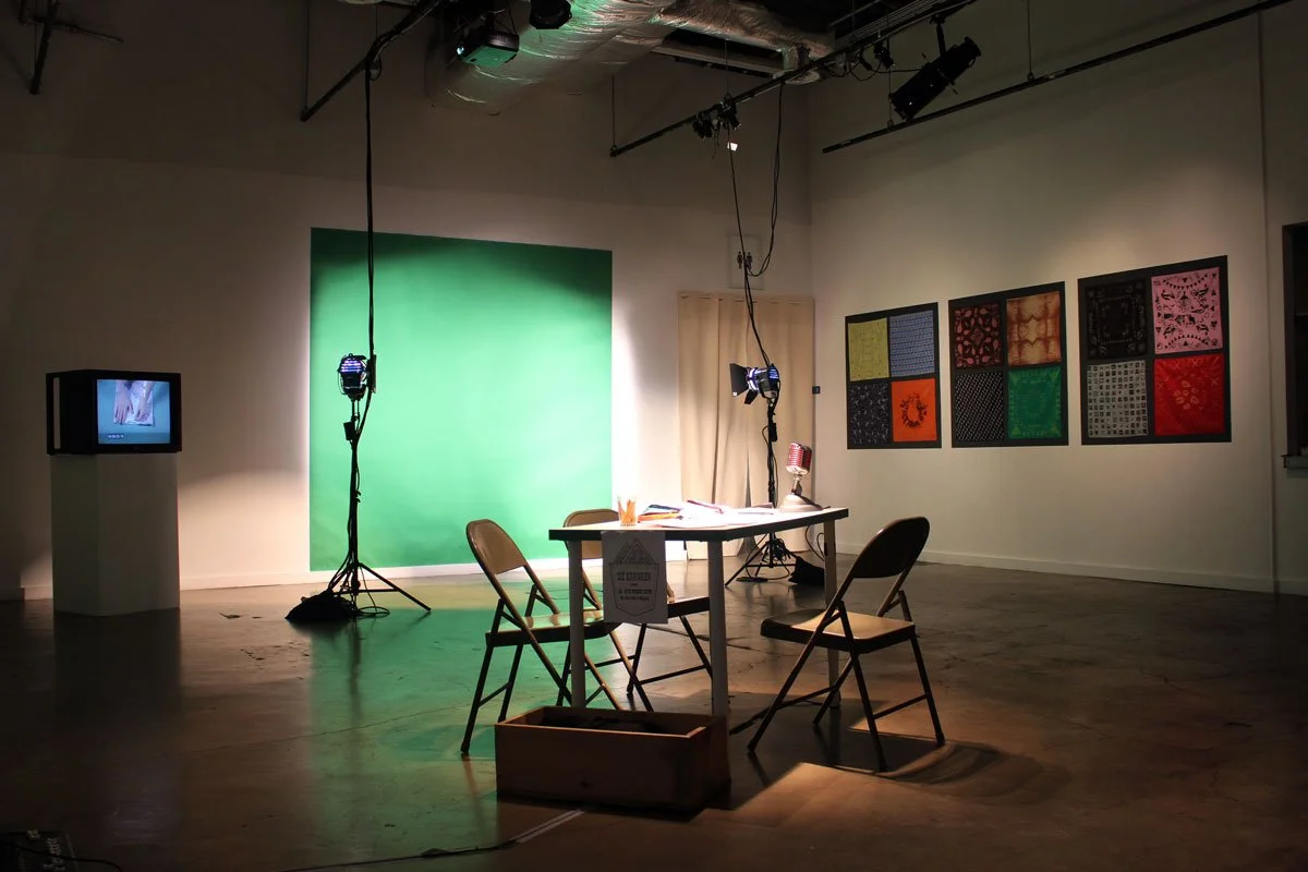 An art studio or gallery setup with a green screen, a small table with two chairs, studio lights, a monitor on a pedestal, and framed textile or fabric art on the walls.