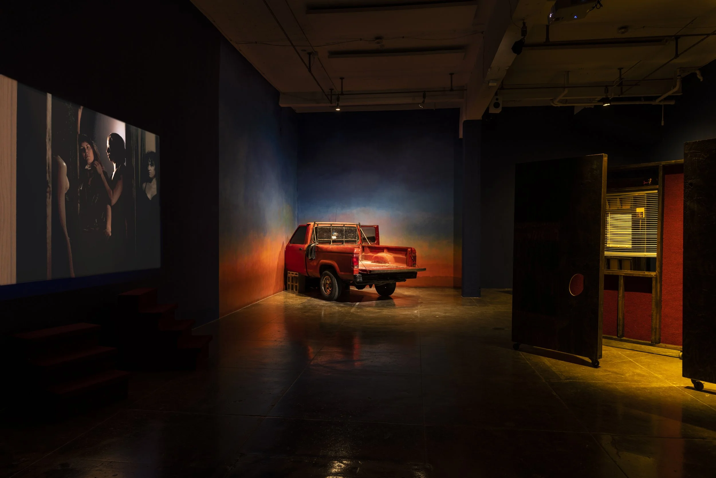 An indoor exhibition space with a red model pickup truck in front of a painted backdrop, and a painting on the left wall showing three women. The space has dark walls and wooden floors.