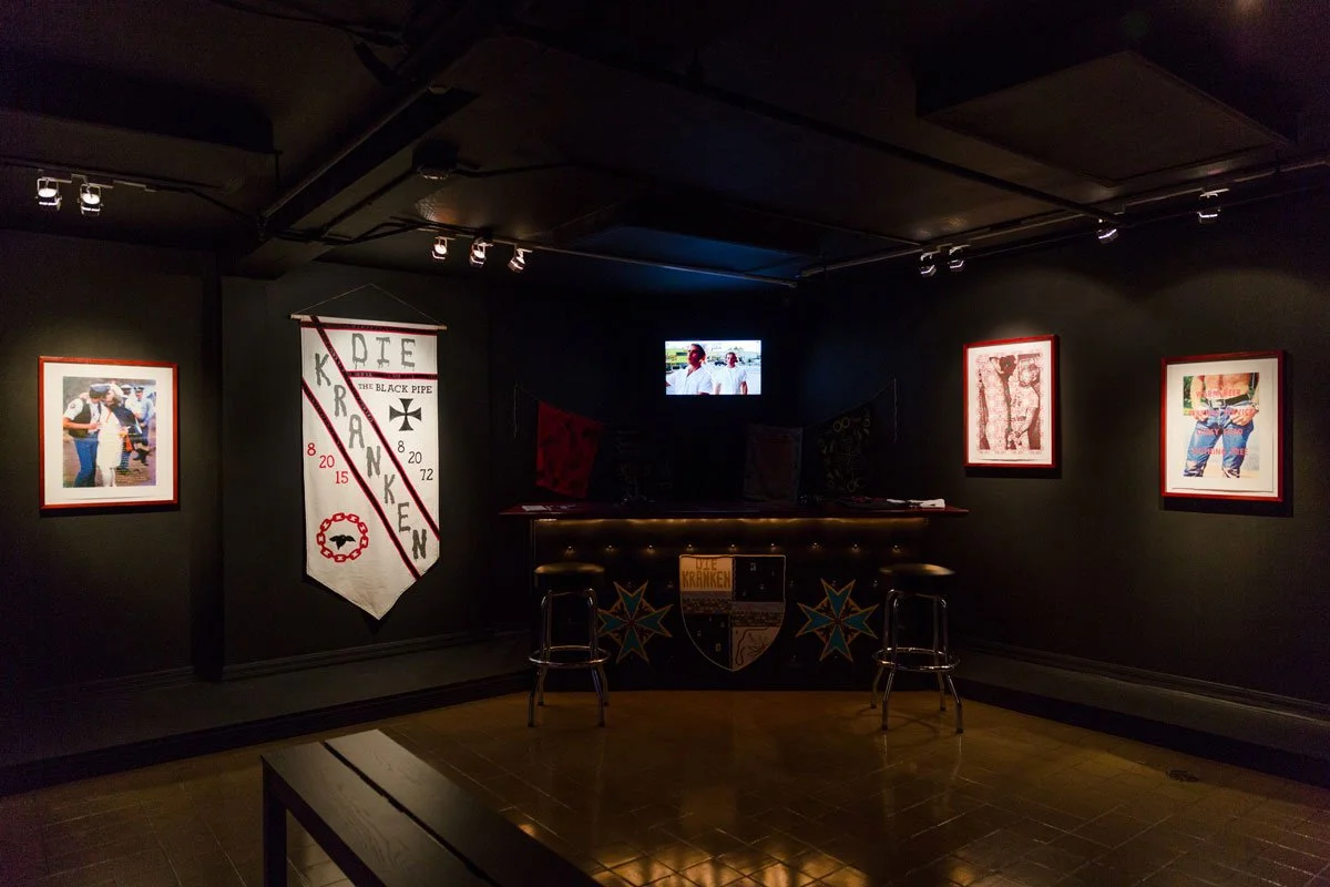 Dimly lit bar with black walls, framed artwork, a white banner with red and black text, and a small television showing two people. There are two bar stools in front of the bar.