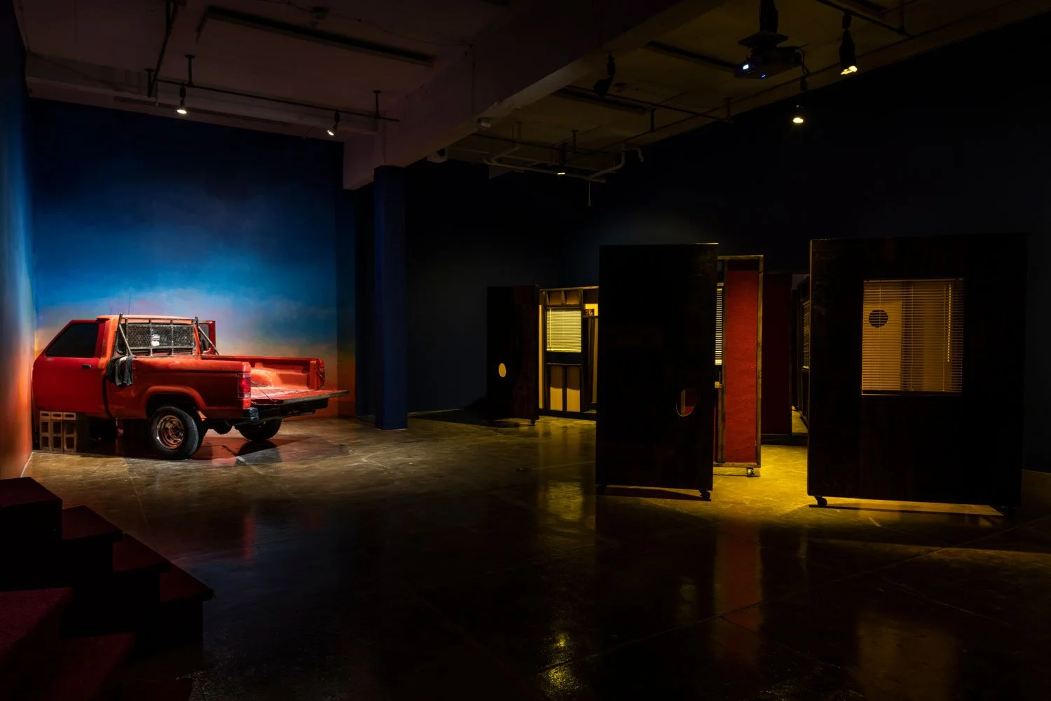 An indoor art installation features a red, vintage-looking pickup truck with a metal mesh barrier in the back, positioned against a colorful gradient wall, with several black and red patterned panels or screens on wheels nearby in a dimly lit space.