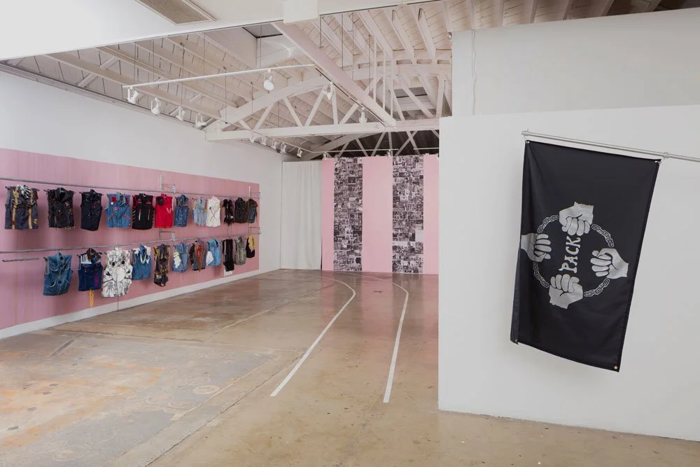 An art gallery with a pink wall displaying various denim outfits and a black flag featuring clenched fists and the word 'BACKS' on a white wall.
