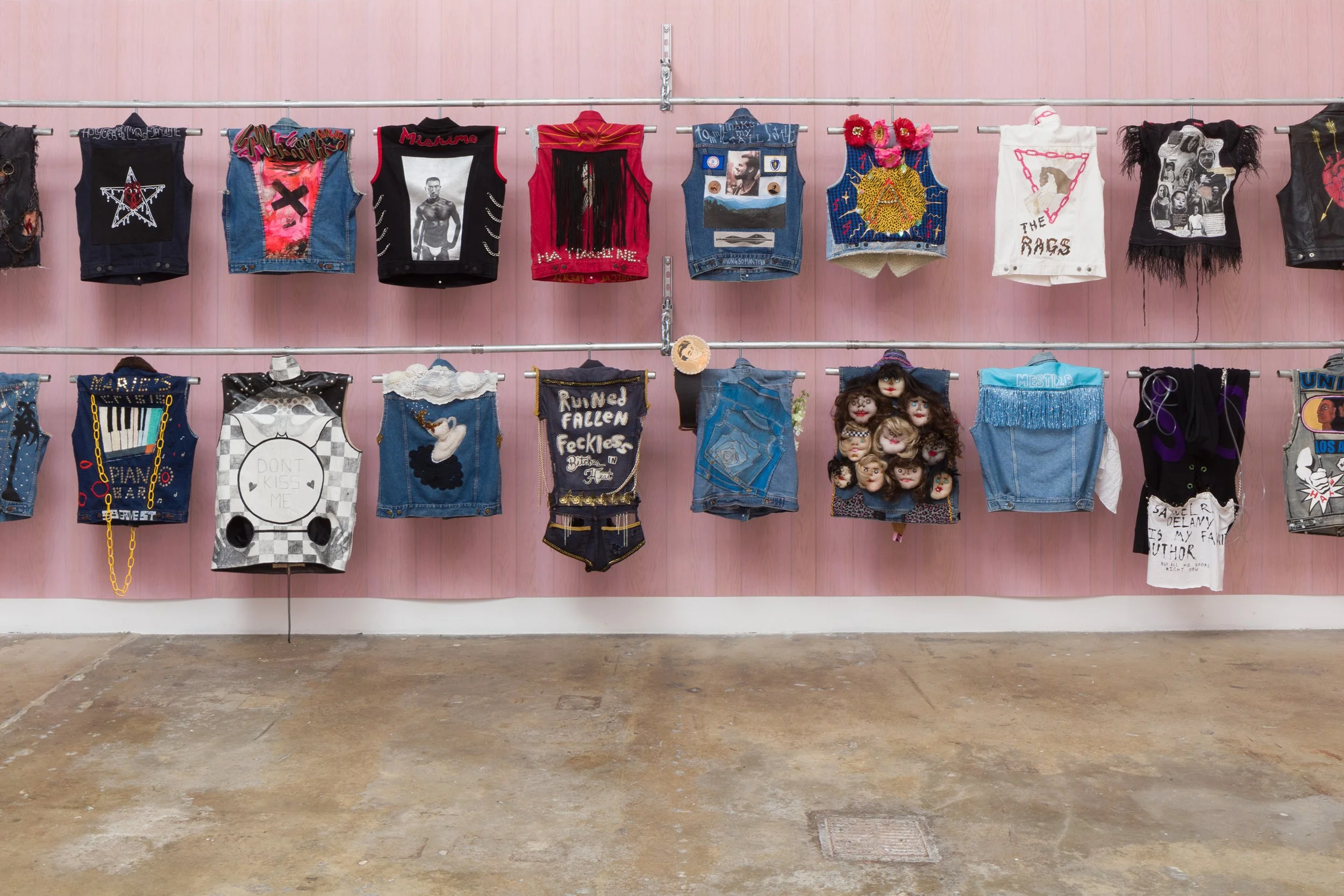 Fashion display with various decorated denim jackets and vests hanging on metal rods against a pink wall.