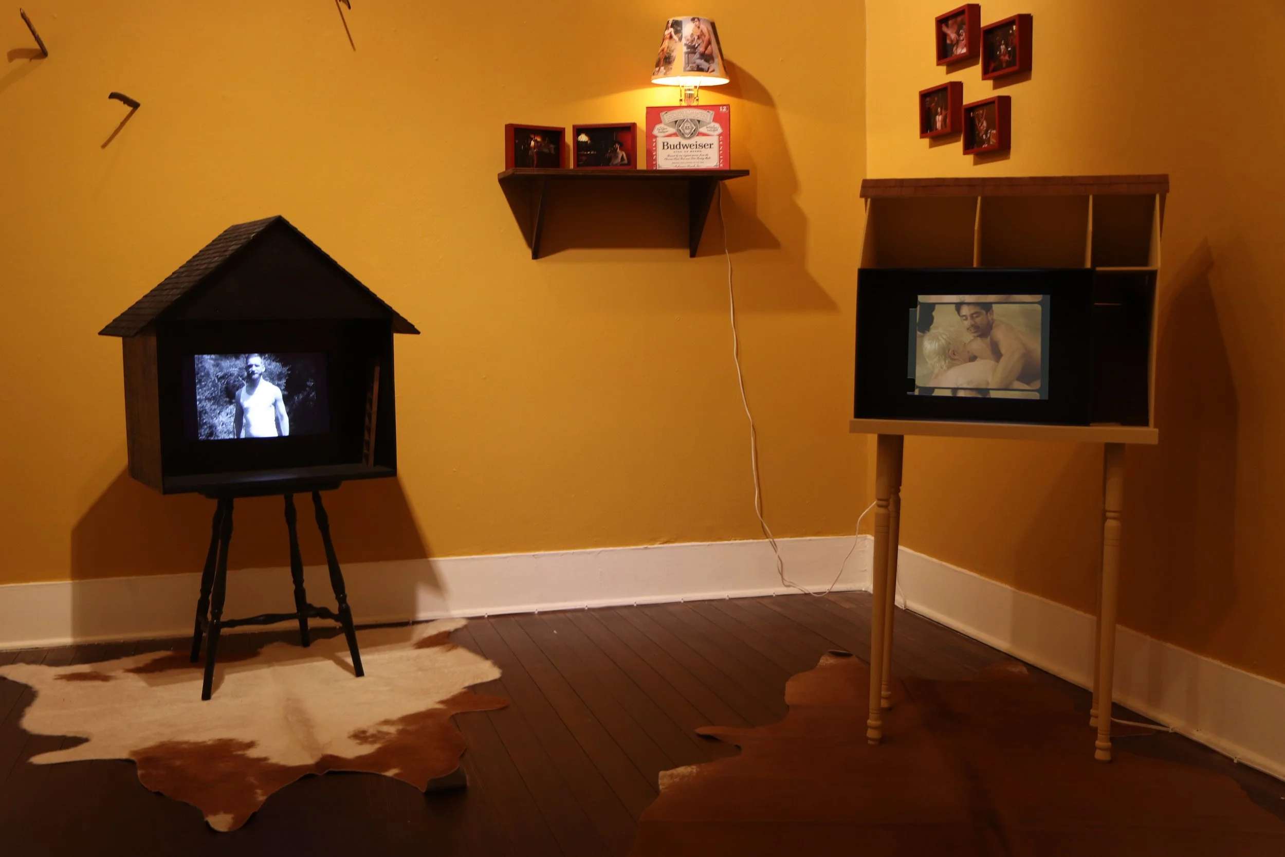 Room with yellow walls, two small TVs on stands, one showing a man shirtless and the other a black-and-white image of a man outdoors, a cowhide rug on the wooden floor, framed photos on the wall, a wooden shelf with framed photos and a Budweiser sign