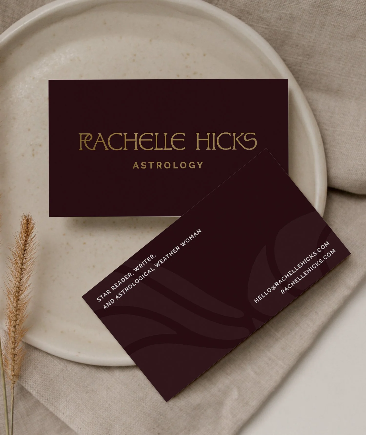 Business card for Rachelle Hicks, an astrology writer and star reader, on a maroon background with gold text, placed on a white ceramic plate and beige fabric. A dried wheat stalk is positioned to the left of the card.