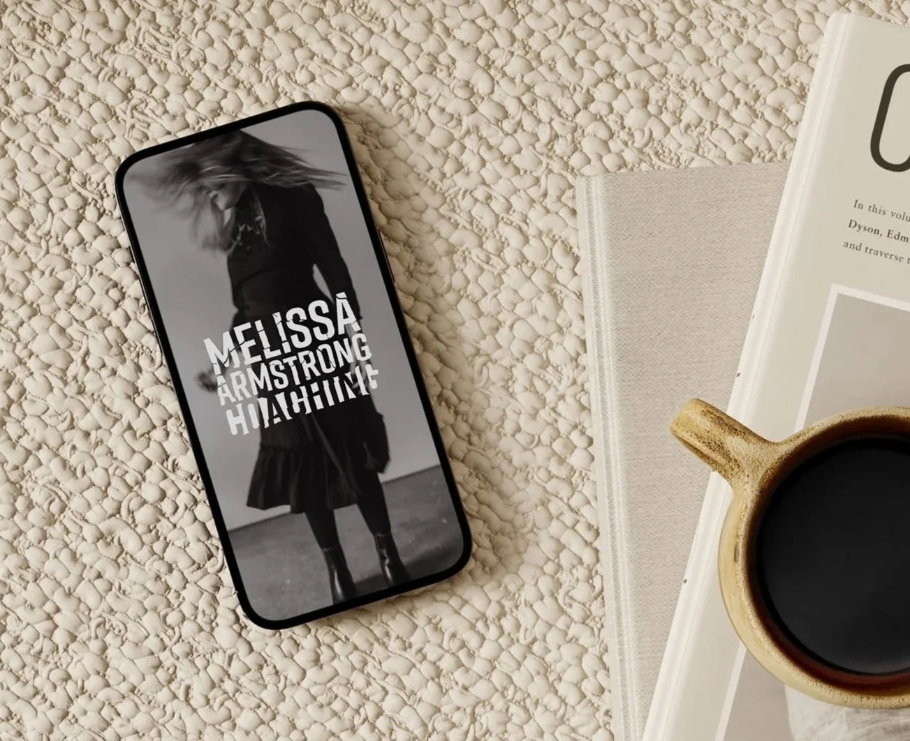 A smartphone displaying a black and white photo of a woman dancing, overlaid with text reading "Melissa Armstrong Hoagime." The phone is on a textured beige surface next to an open book, a beige cloth, a cup of black coffee, and a plate.