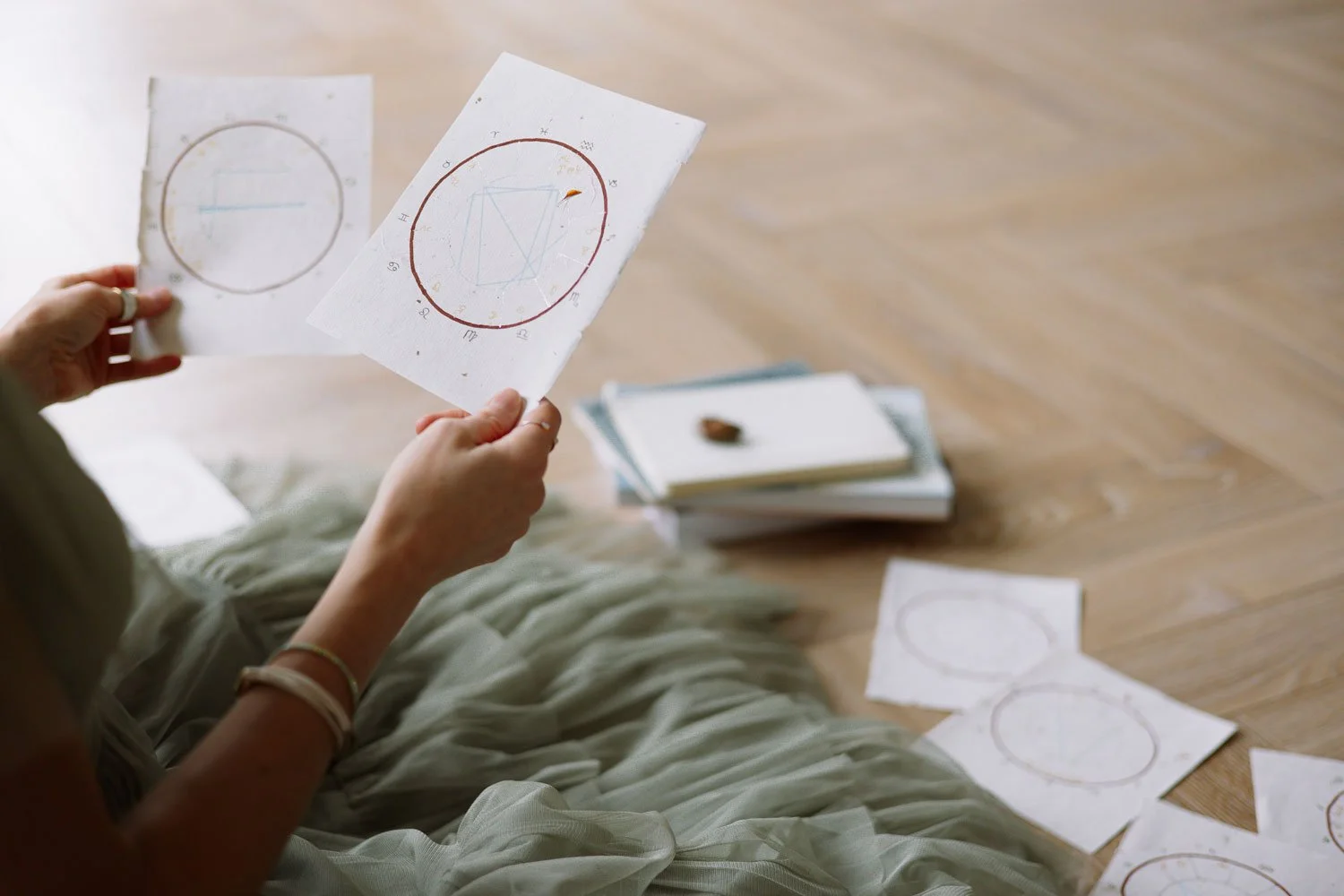 Rachelle Hicks of Rachelle Hicks Astrology holding printed natal chart wheels on a wood floor surrounded by additional birth chart printouts and stacked notebooks