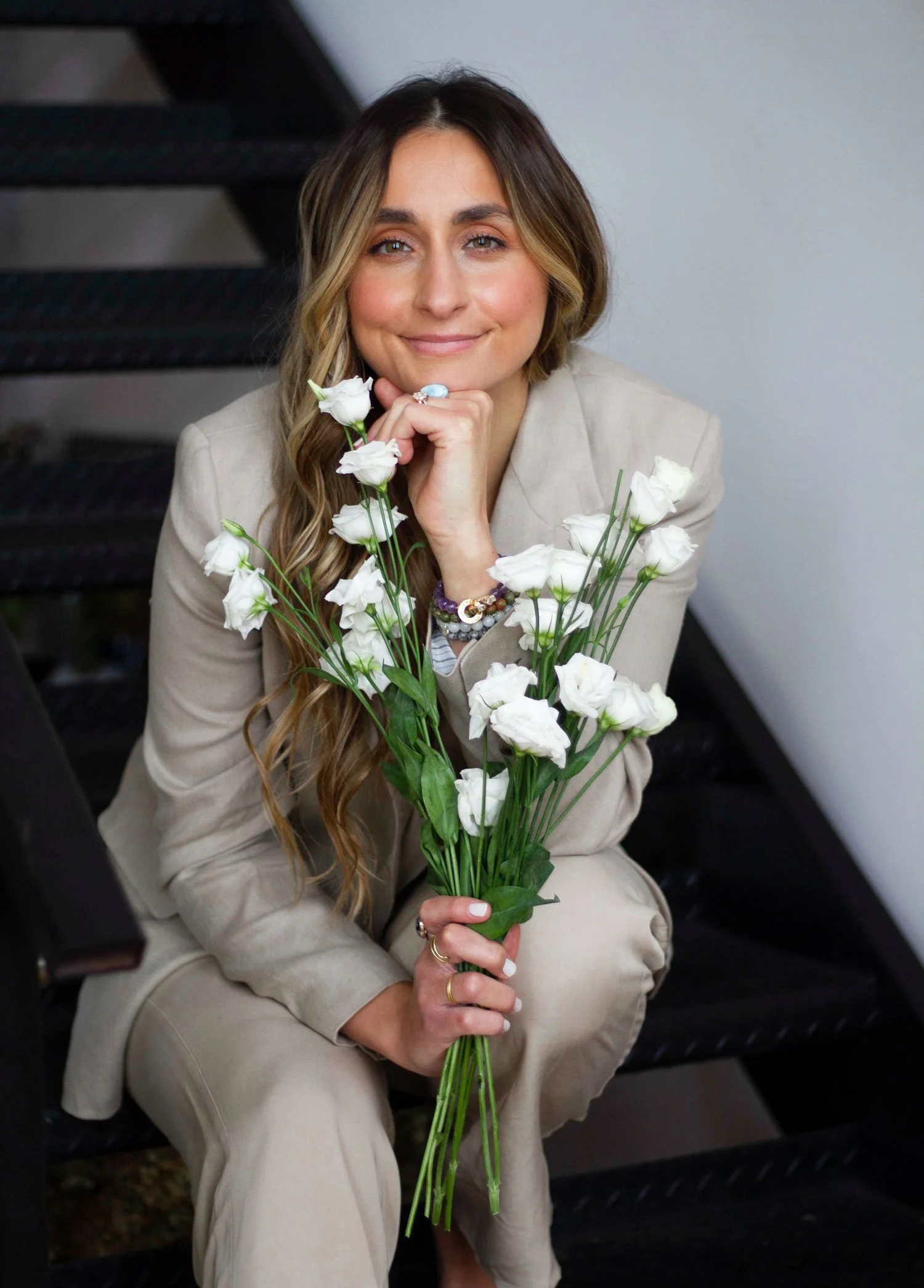Natalie Kehren seated on stairs in a neutral suit holding white roses