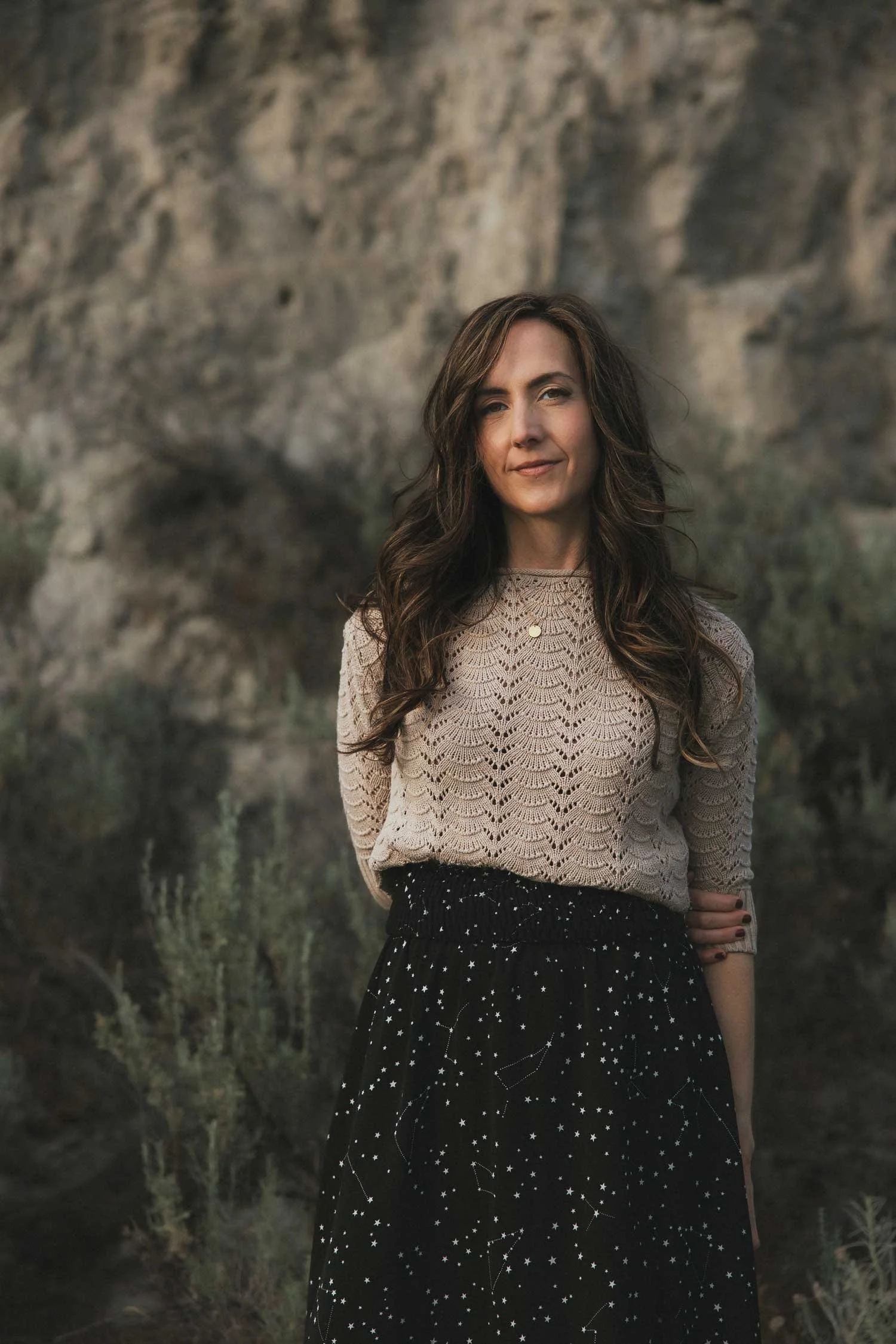 Moonstone Creative founder, Miranda Braithwaite, standing outdoors in a cream sweater and black constellation skirt against a rocky Okanagan landscape