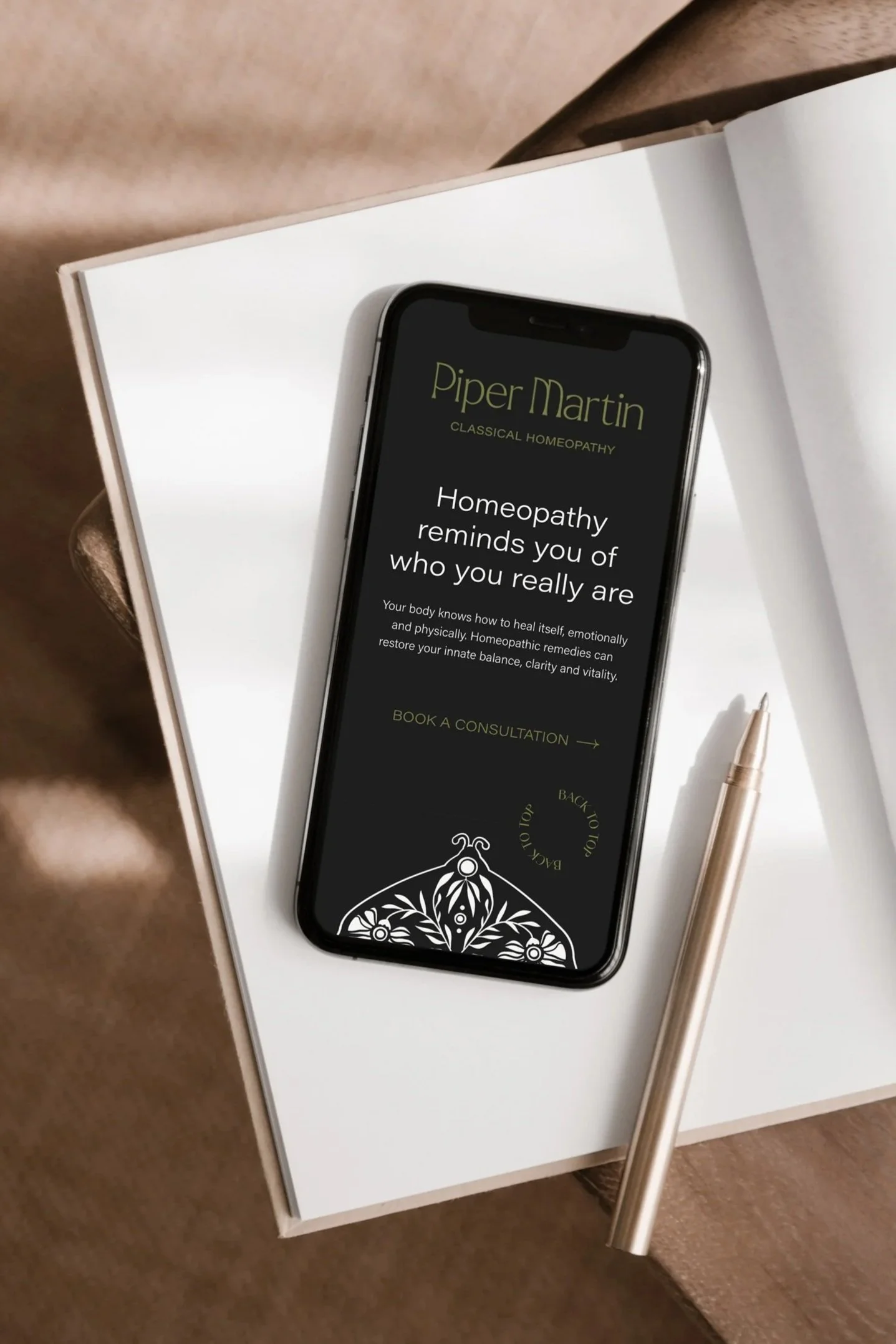 A smartphone displaying a website for Piper Martin Classical Homeopathy, placed on a white open notebook with a gold pen next to it, on a brown surface.