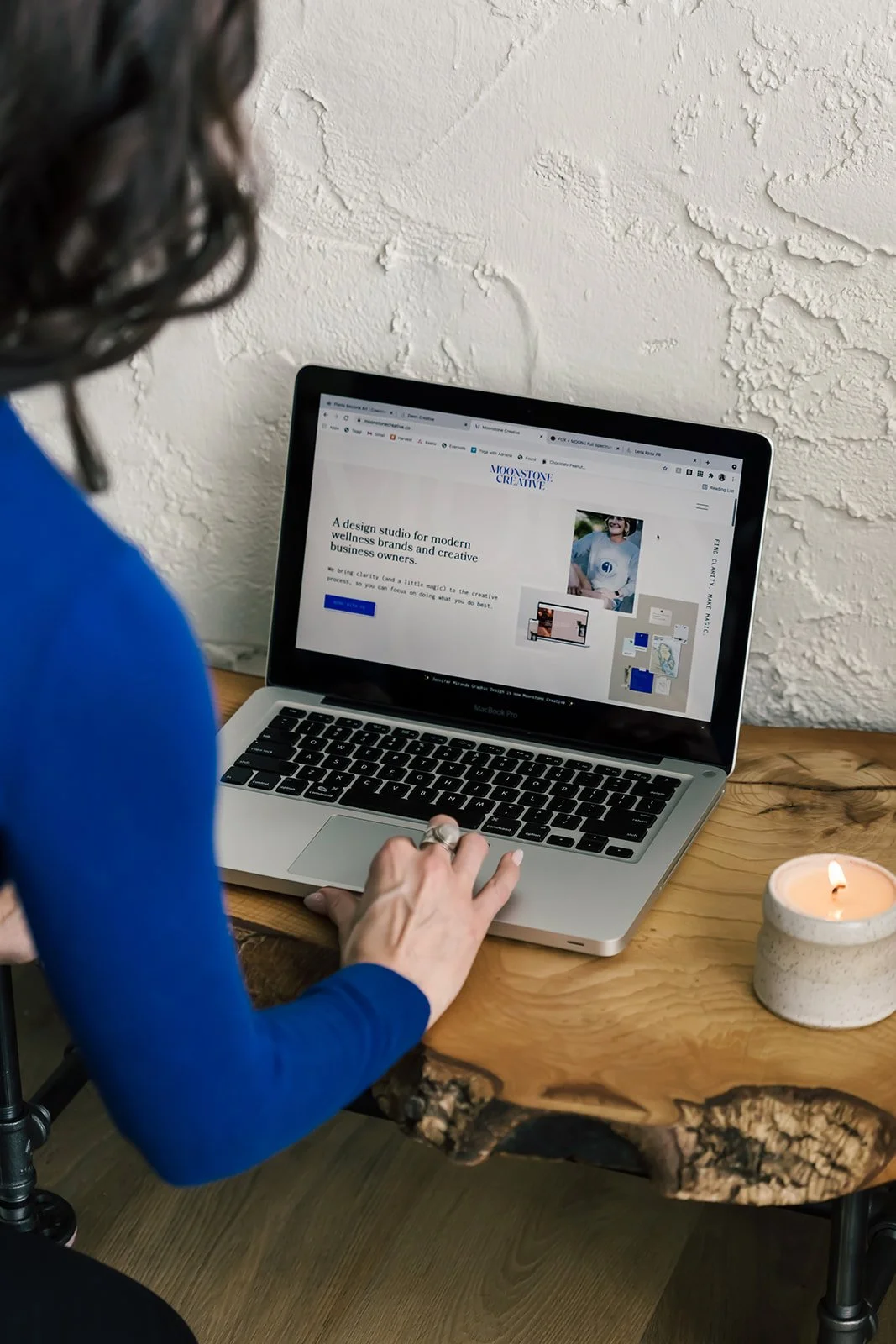 Miranda wearing an electric blue top while working on her MacBook Pro at a wooden desk with a lit candle nearby, representing a brand colour palette that looked beautiful but didn't feel personally aligned.