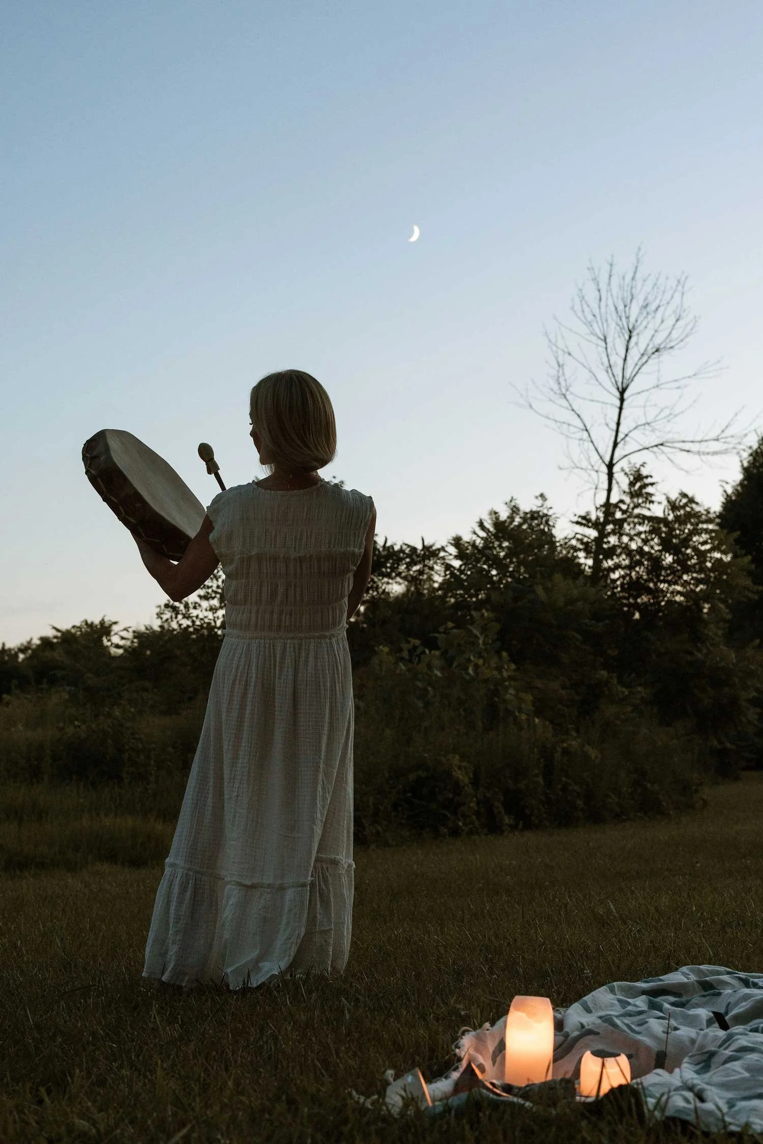 Ashley Livingston of Live As The Light playing a frame drum outdoors at dusk beneath a crescent moon with a glowing candle on a blanket nearby