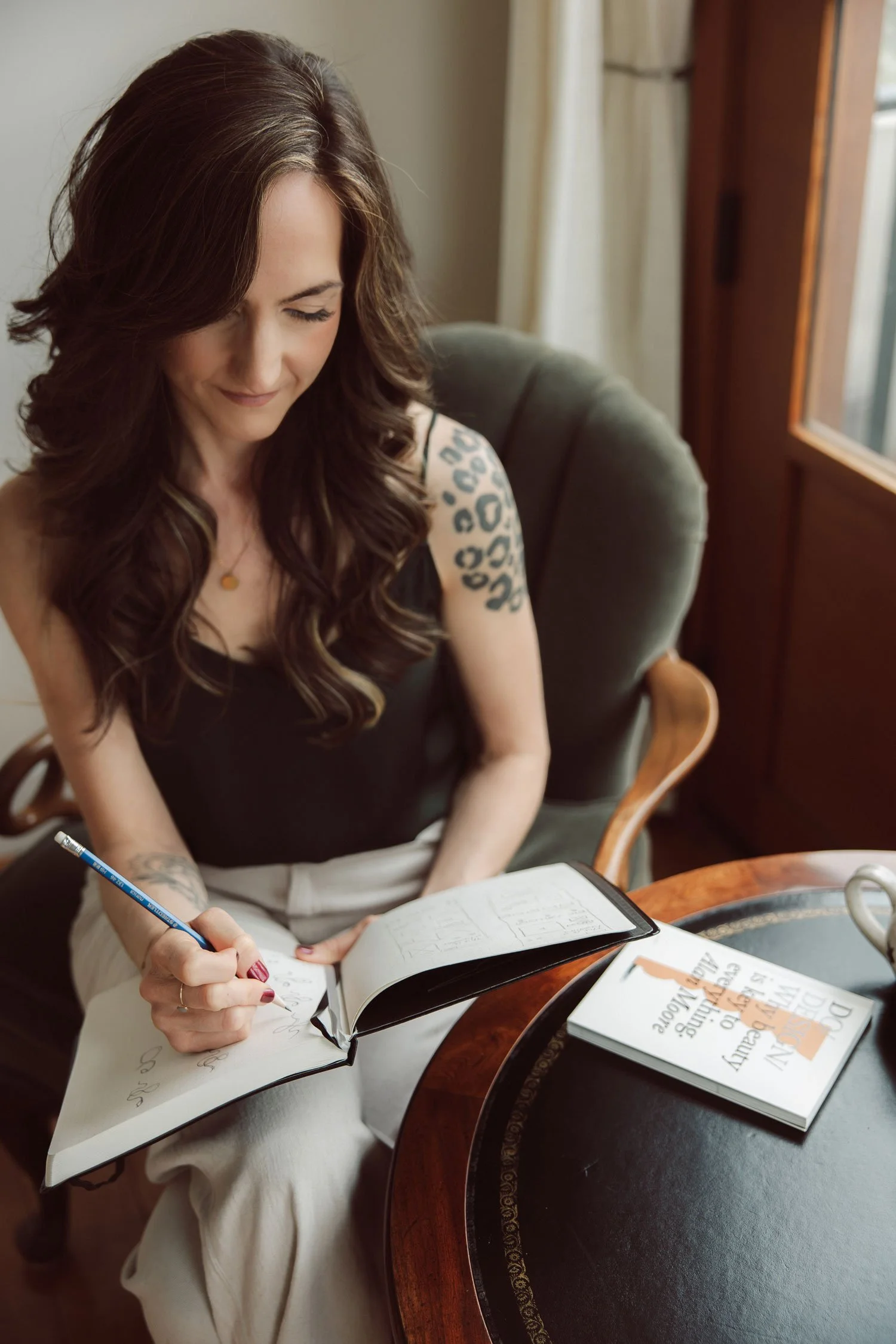 Miranda Braithwaite, founder of Moonstone Creative, sketching brand layouts in a notebook beside an antique side table with a design book nearby