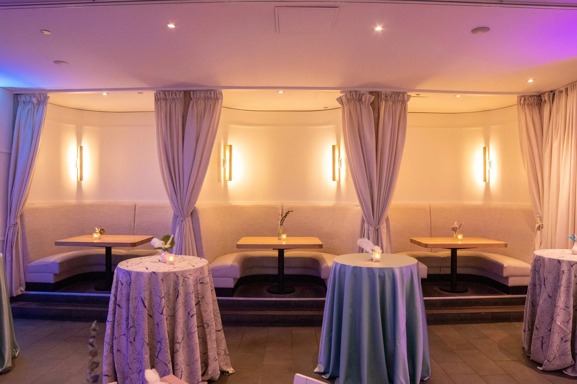 Elegant indoor dining area with curved seating, beige cushions, beige curtains, and soft lighting, featuring small tables with candles and flowers.