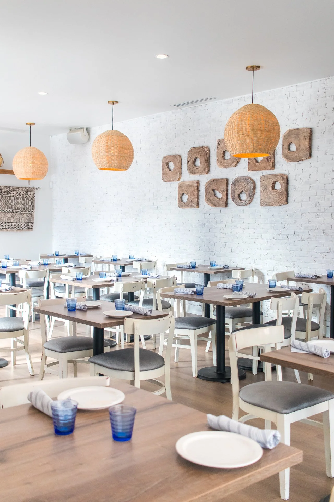 Empty restaurant dining area with white tables and chairs, blue glasses, and white plates, decorated with woven pendant lights and wall art.