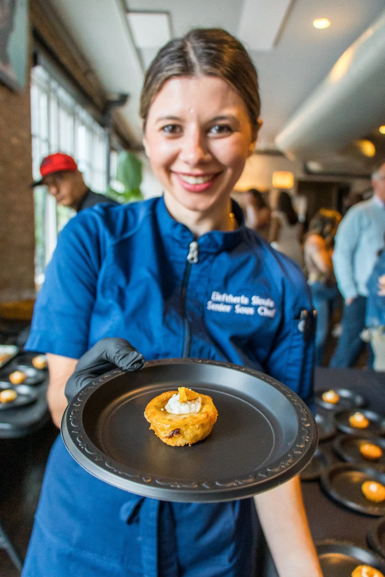 A smiling woman wearing a blue jacket holding a plate with a baked appetizer topped with cream and an orange garnish, at a food event.