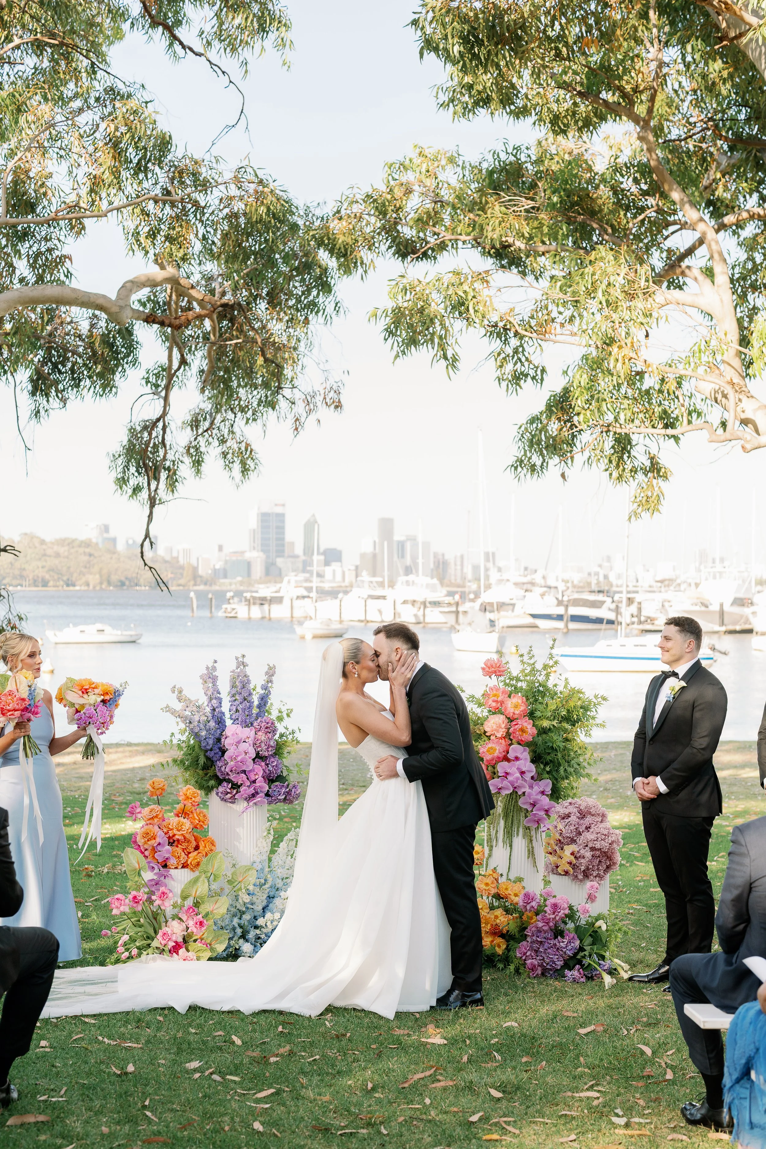 A colourful wedding ceremony at Matilda Bay Restaurant, on the banks of the swan river in Perth, capture by Perth Wedding Photographer Natalie Jane. Colourful florals by Perth wedding Florist, The Flower Motel, created the perfect arbour.