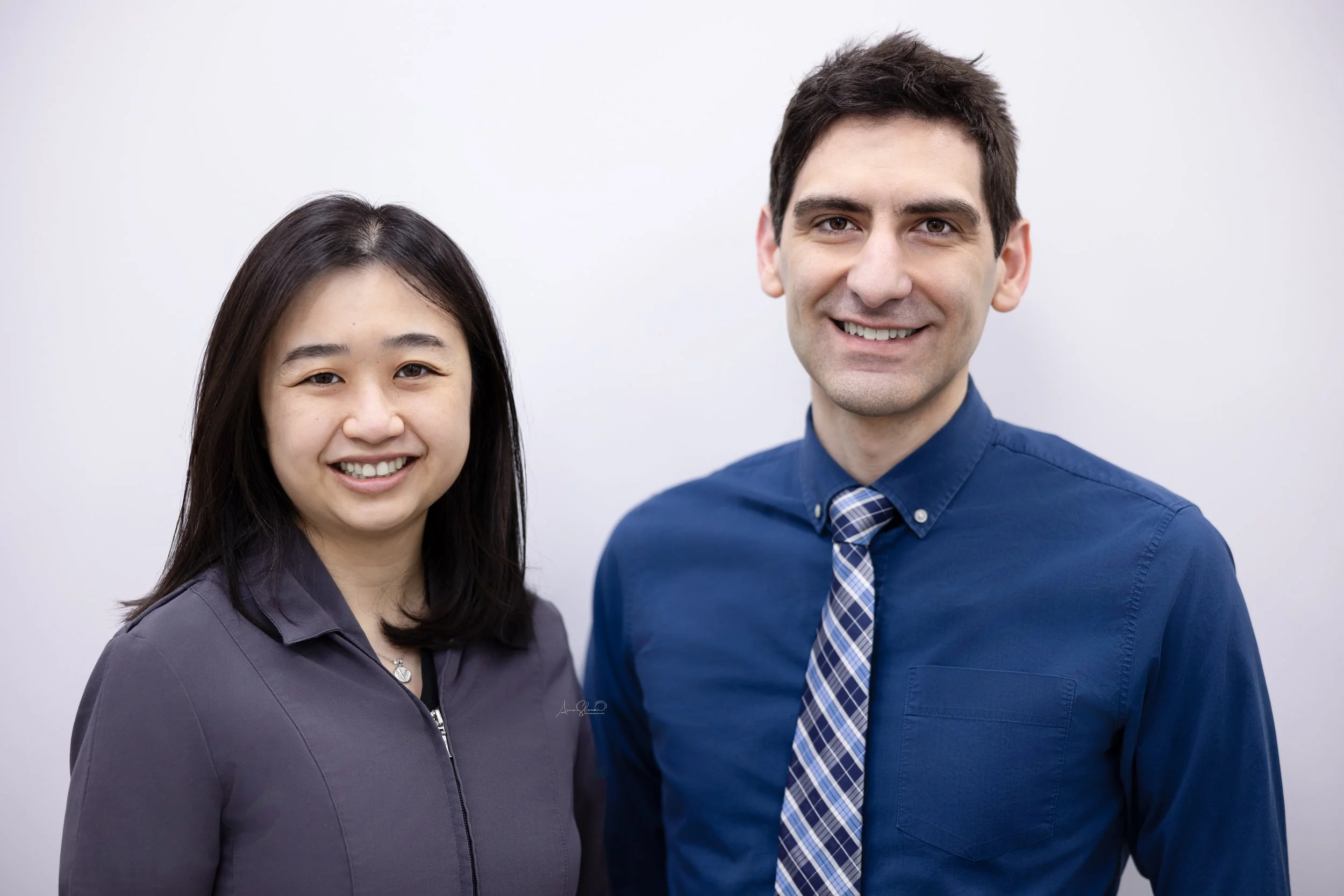 Two smiling professionals, a woman of Asian descent in a gray blazer and a man of Caucasian descent in a blue shirt with a plaid tie, standing side by side against a white background.