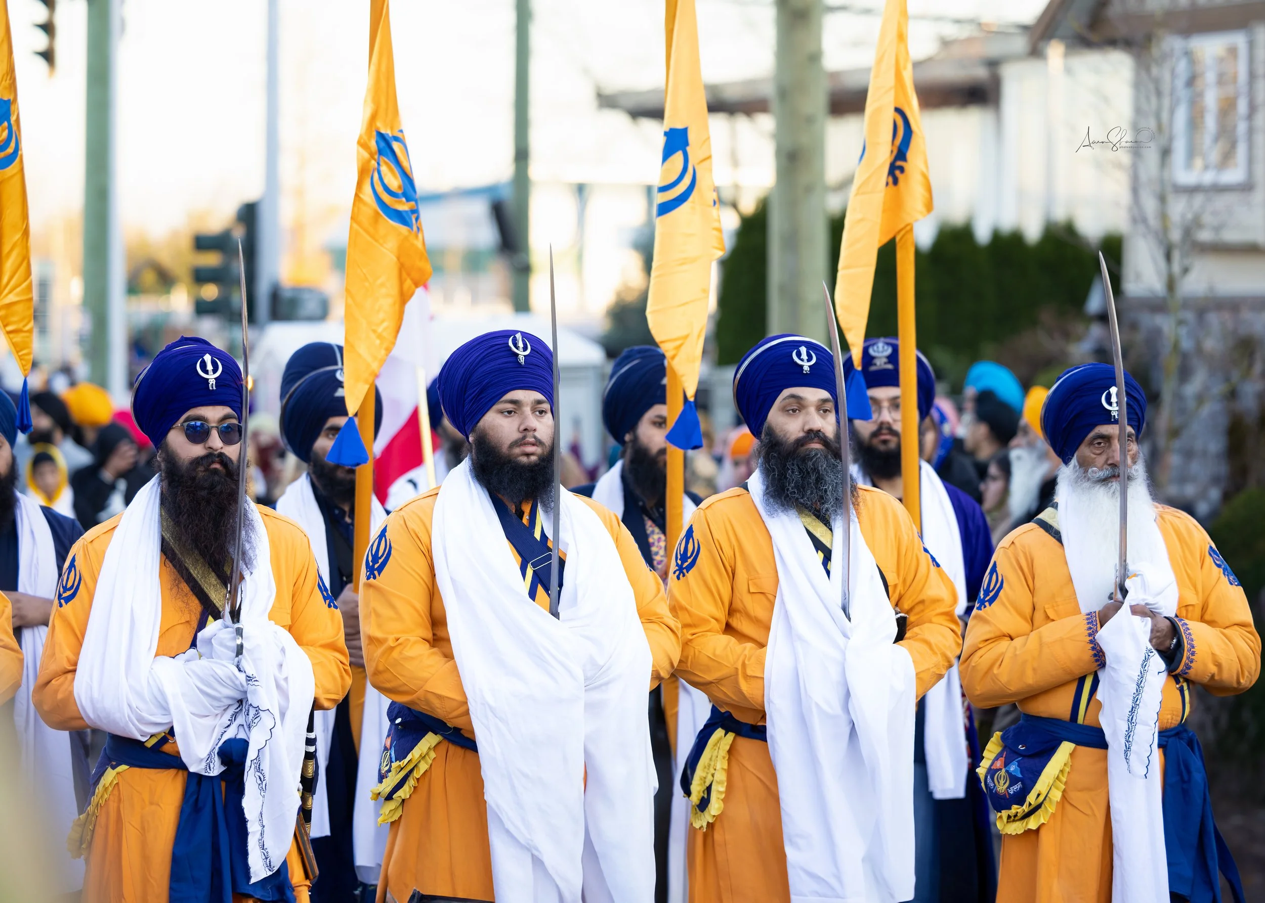 Group of Sikh men dressed in traditional orange and blue attire, holding yellow flags with blue symbols, participating in a parade or gathering.