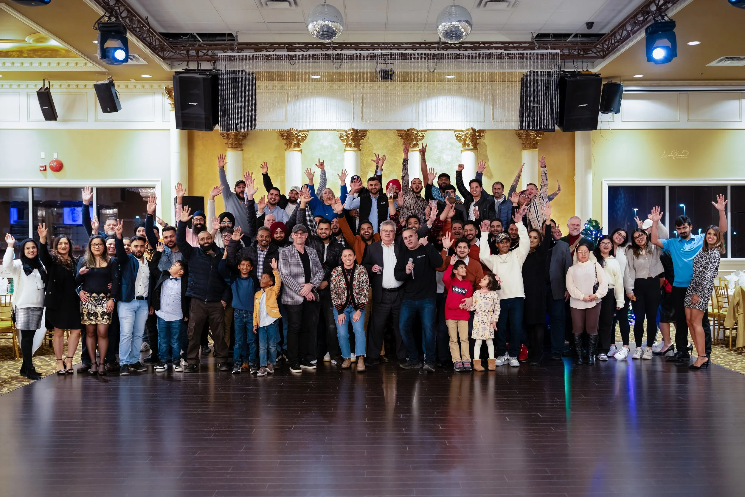 Large group of people gathered in a banquet hall, smiling and raising their hands for a group photo. The hall is decorated with gold and white accents, with a dark wood floor and stage area.