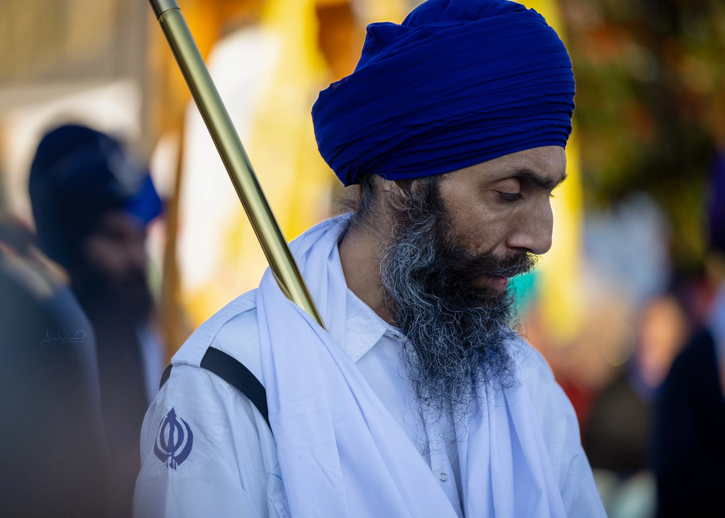 A man wearing a blue turban and white clothing with a religious symbol, standing outdoors with his eyes closed, holding a staff on his shoulder, in a contemplative moment surrounded by blurred colorful background.