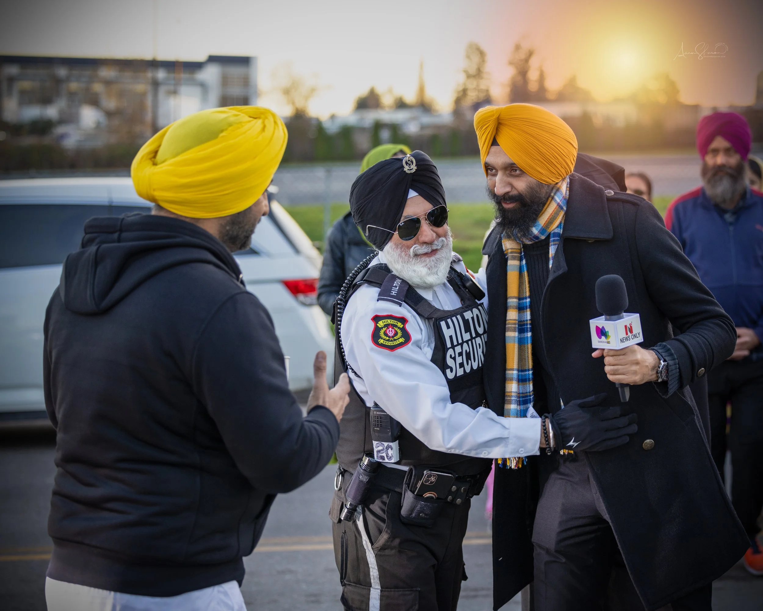 A Sikh man holding a microphone, interviewing a man wearing a yellow turban, who is greeting another man in a black coat with a yellow turban during a gathering outdoors at sunset with several other people in the background.