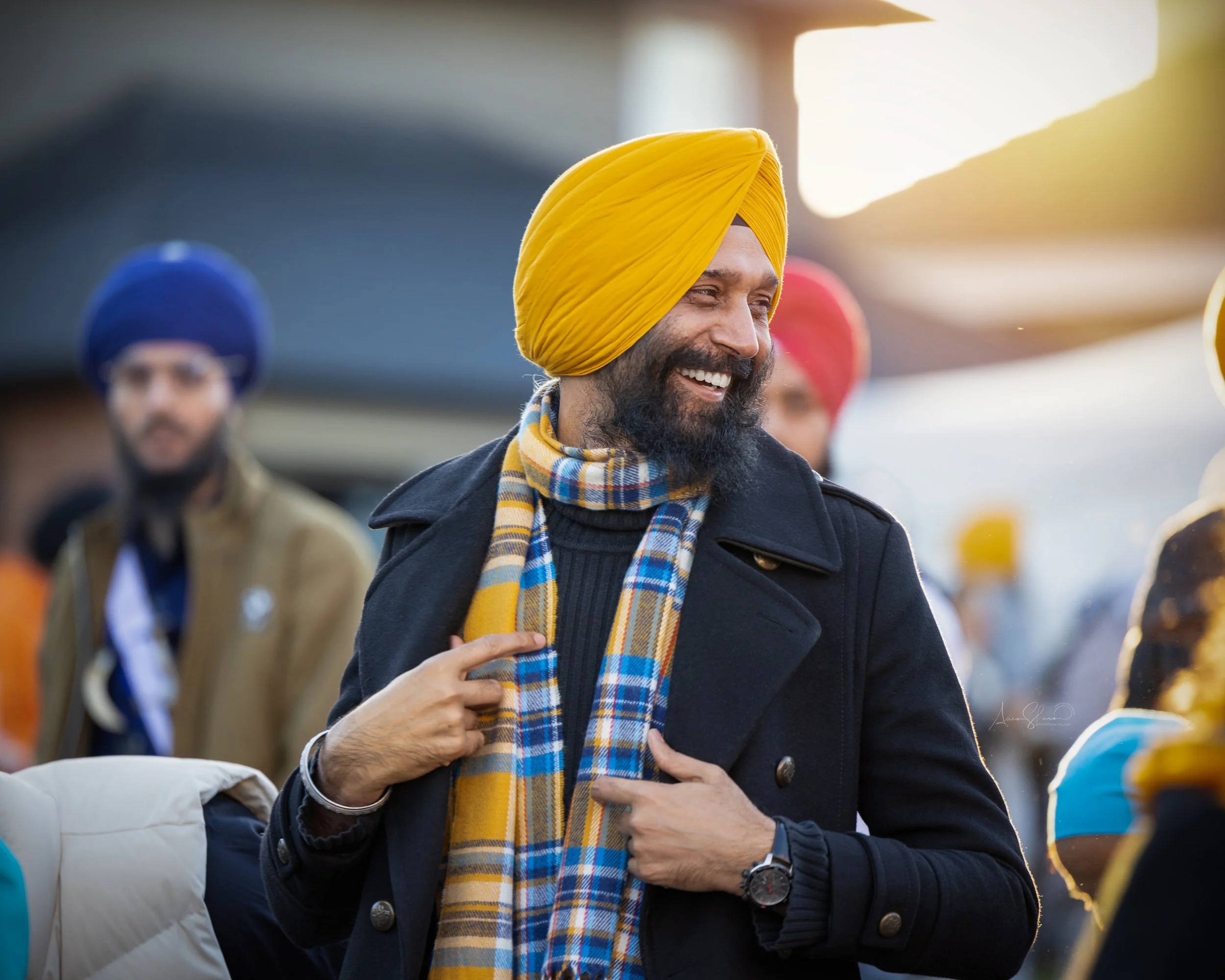 A man with a yellow turban smiling and pointing to himself at an outdoor event, with other people wearing turbans in the background.