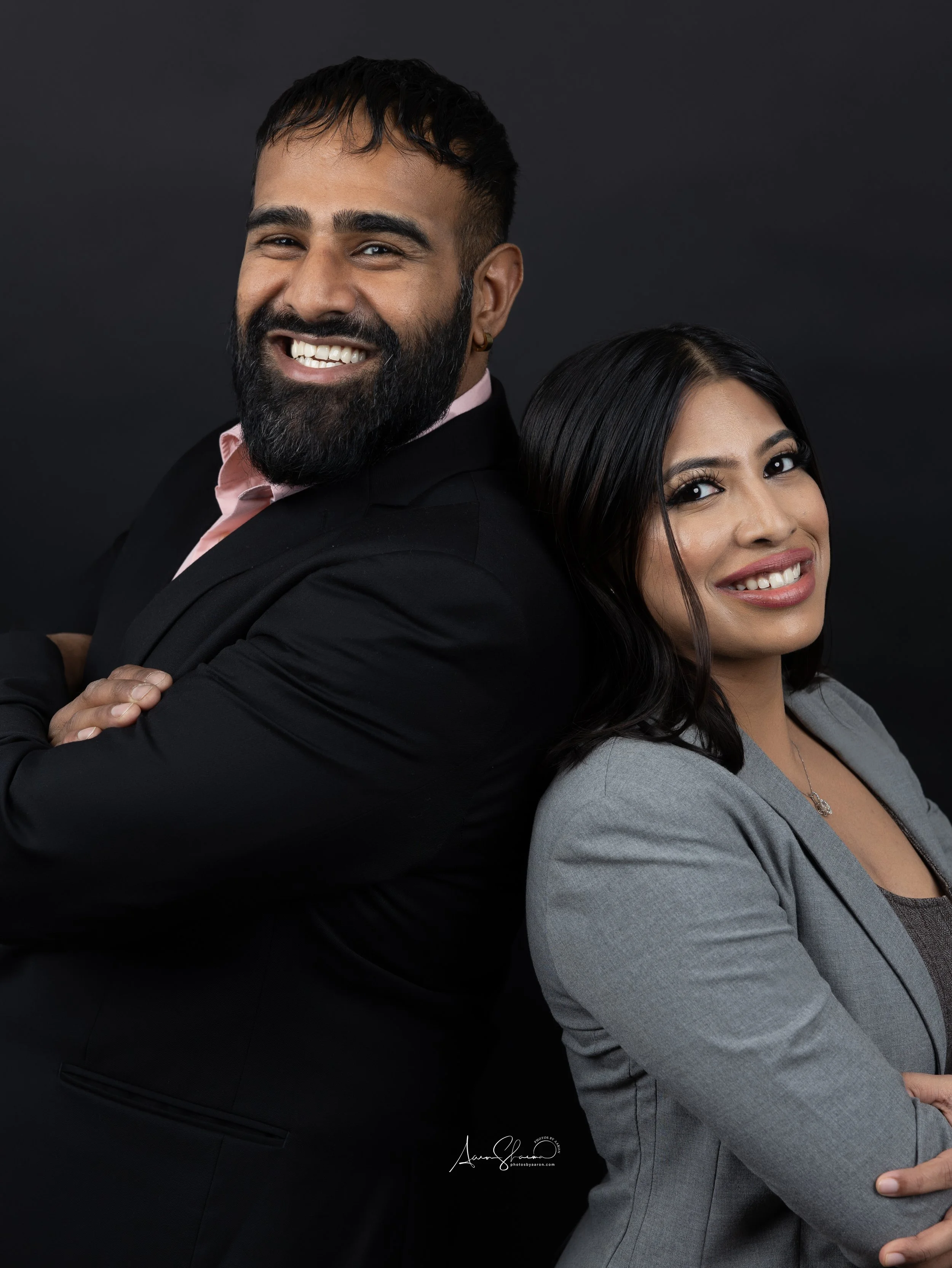 A smiling man with a beard and a woman with dark hair, both dressed in business attire, standing back-to-back against a dark background.
