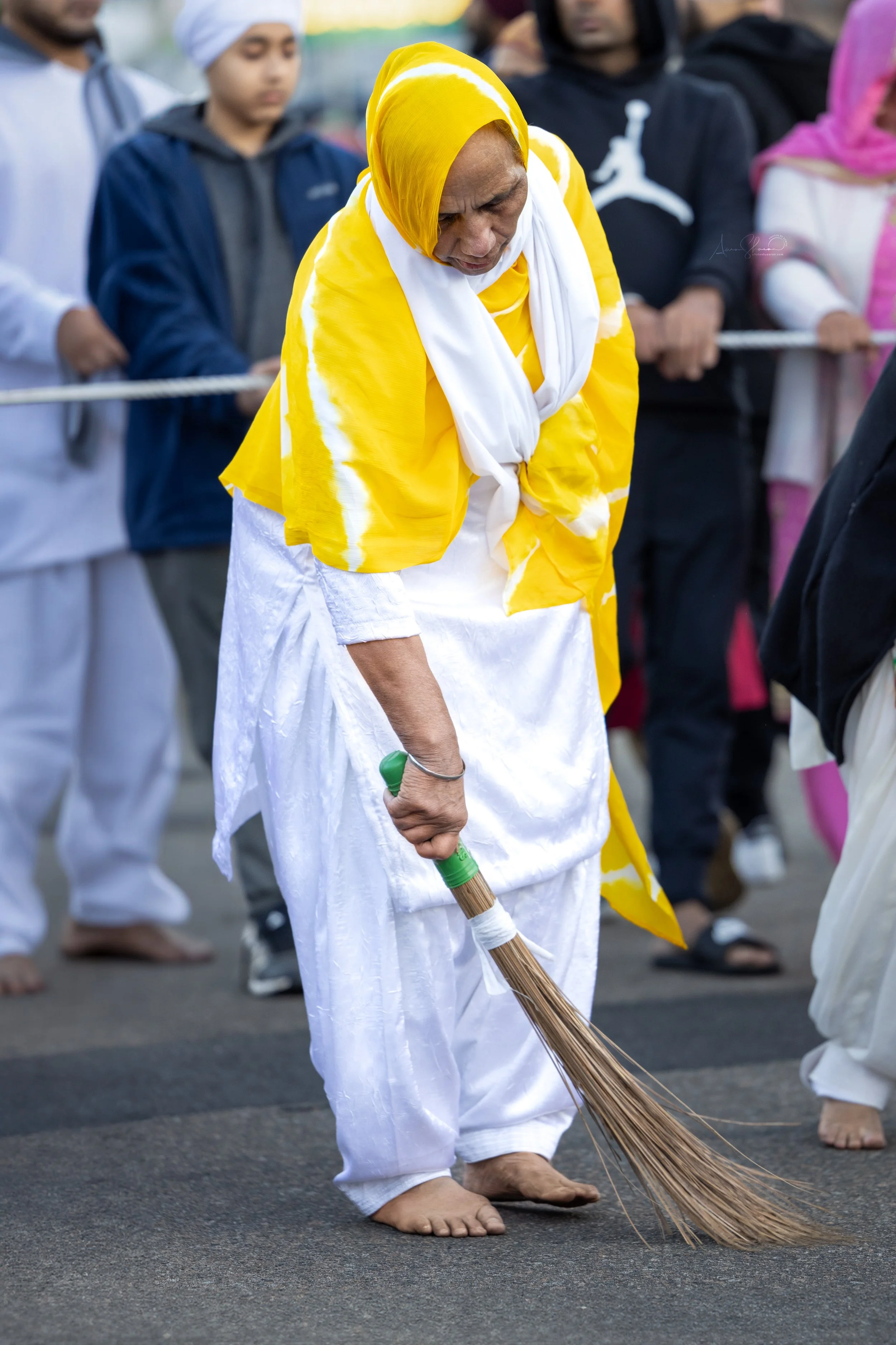 An elderly woman dressed in traditional Indian clothing, including a yellow and white shawl, sweeping the ground with a broom during a public event or procession surrounded by a group of people.