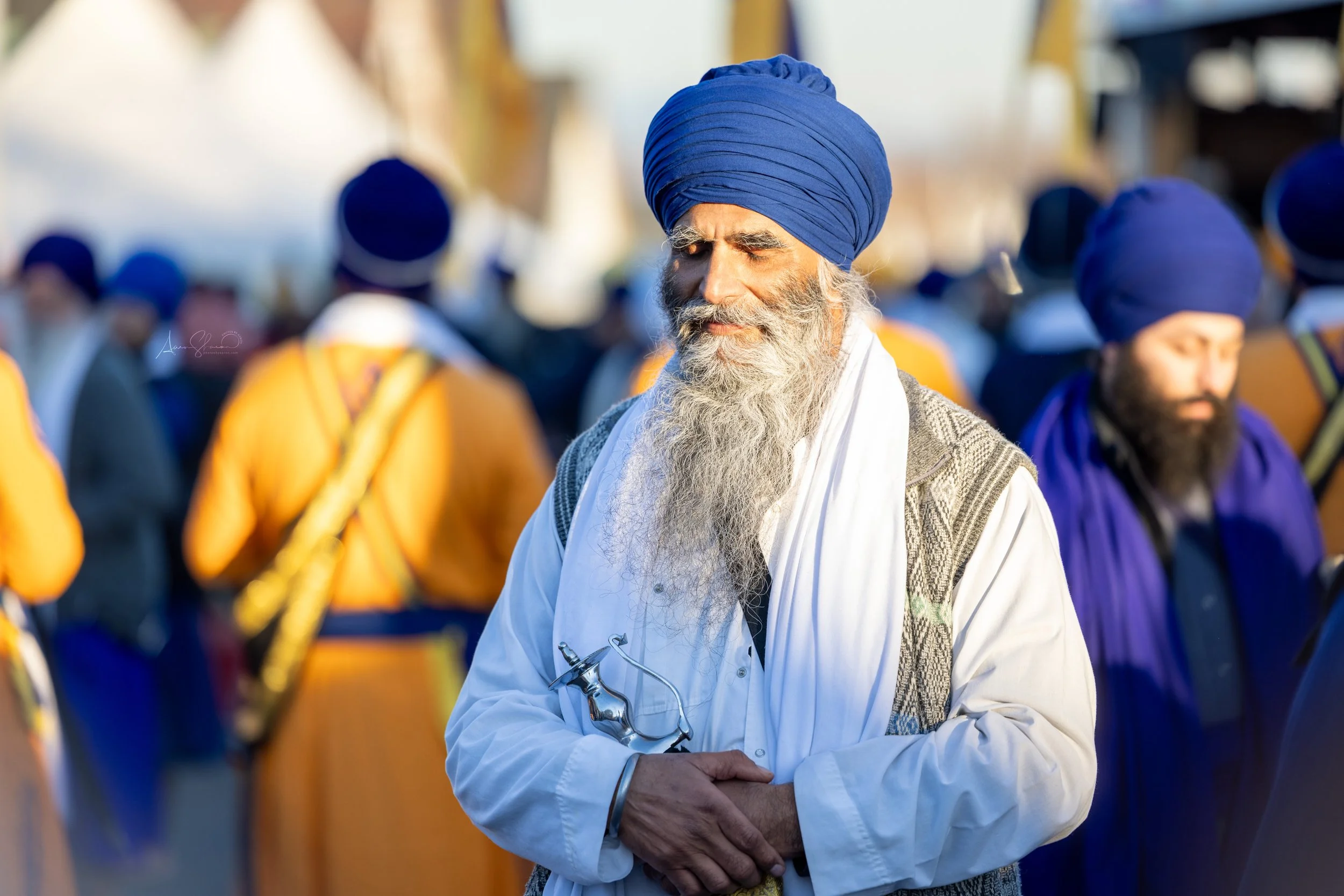 An elderly man in a blue turban and white traditional clothing standing among a crowd of men wearing turbans and colorful clothing, outdoors during the daytime.