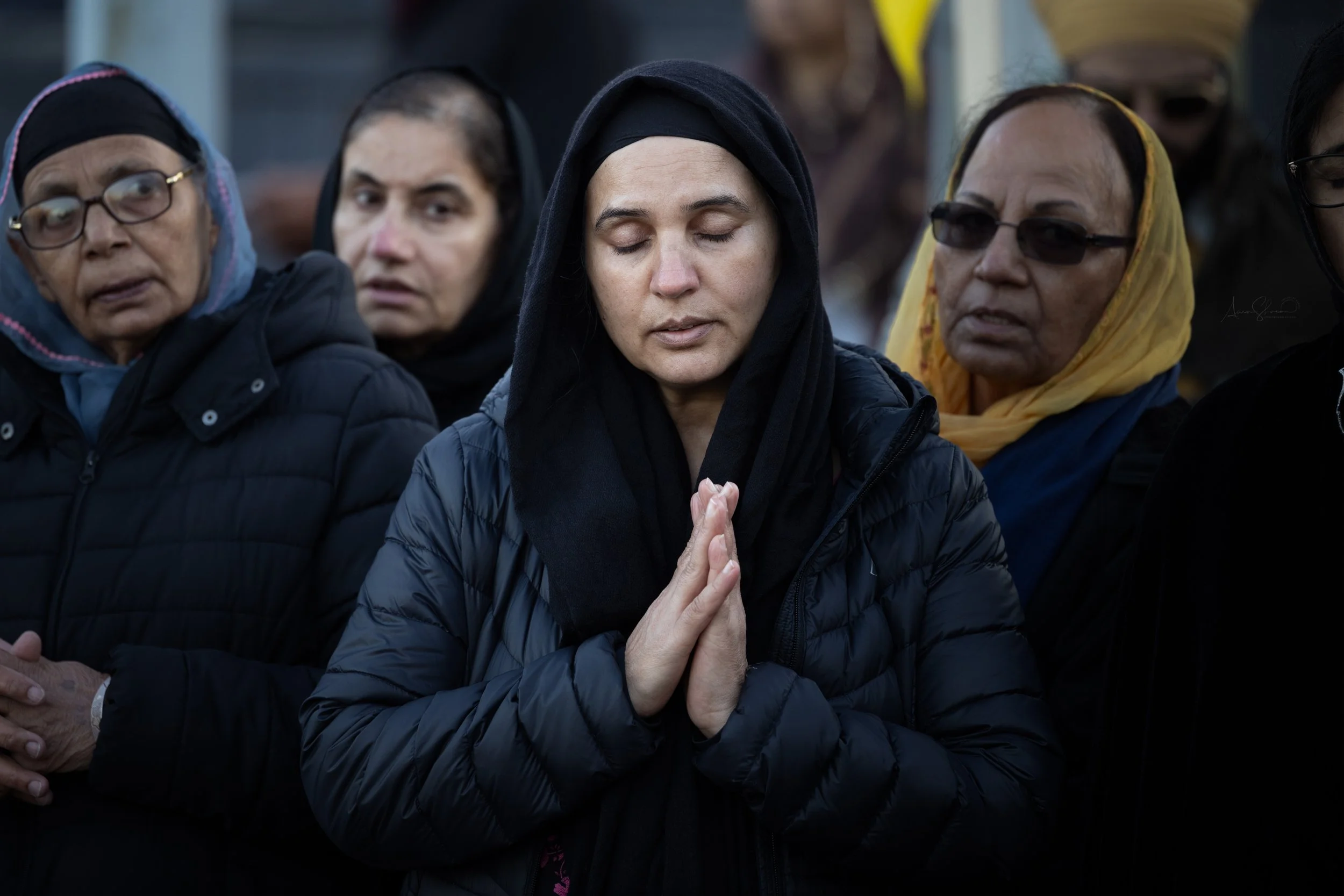 A group of women with somber expressions, some with eyes closed and hands clasped in prayer, participating in a somber outdoor event.