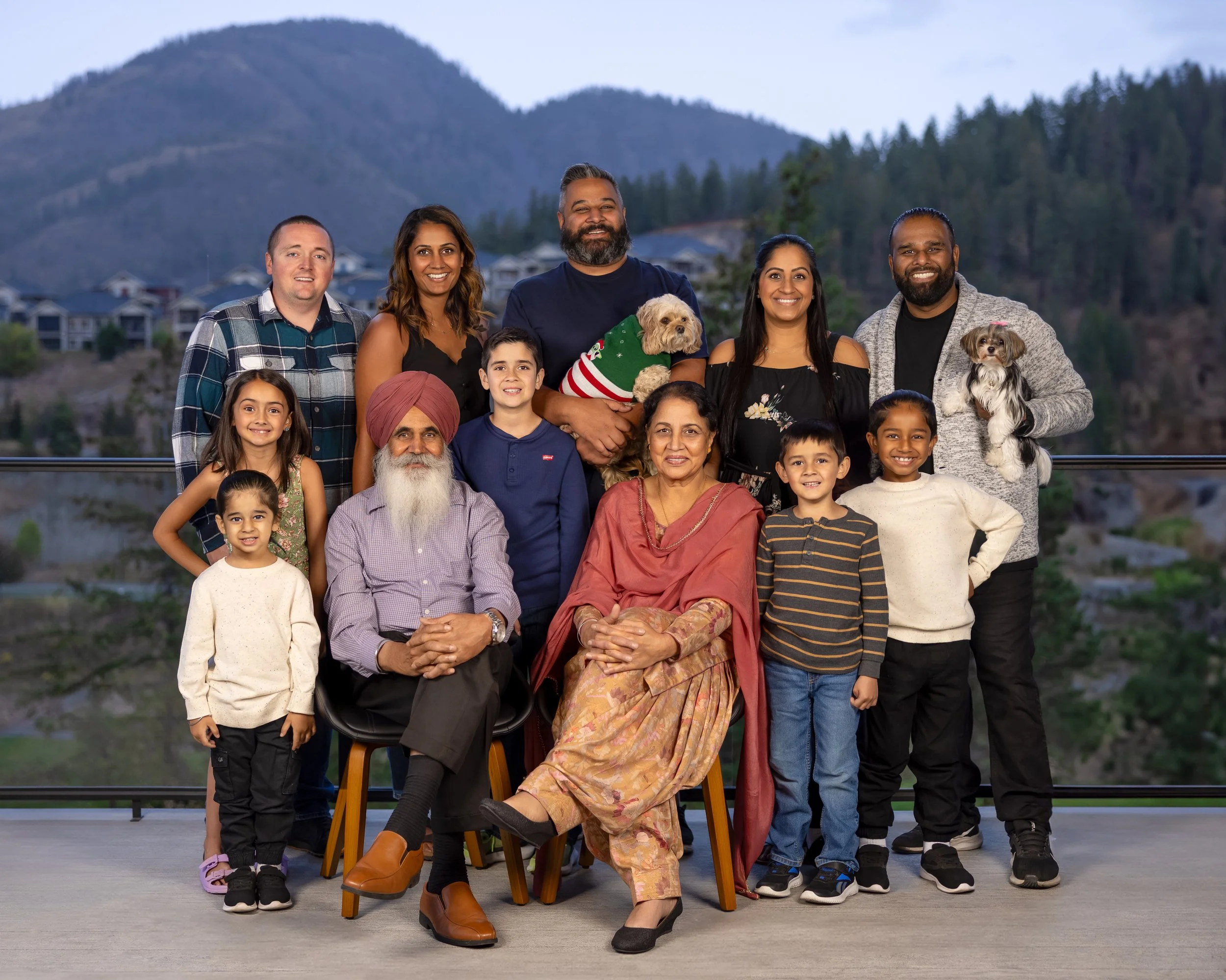 Black and white photo of multiple generations of a family, including children, adults, and elderly, gathered together on a balcony with mountain scenery in the background. Some are holding dogs, and everyone is smiling and looking at each other.