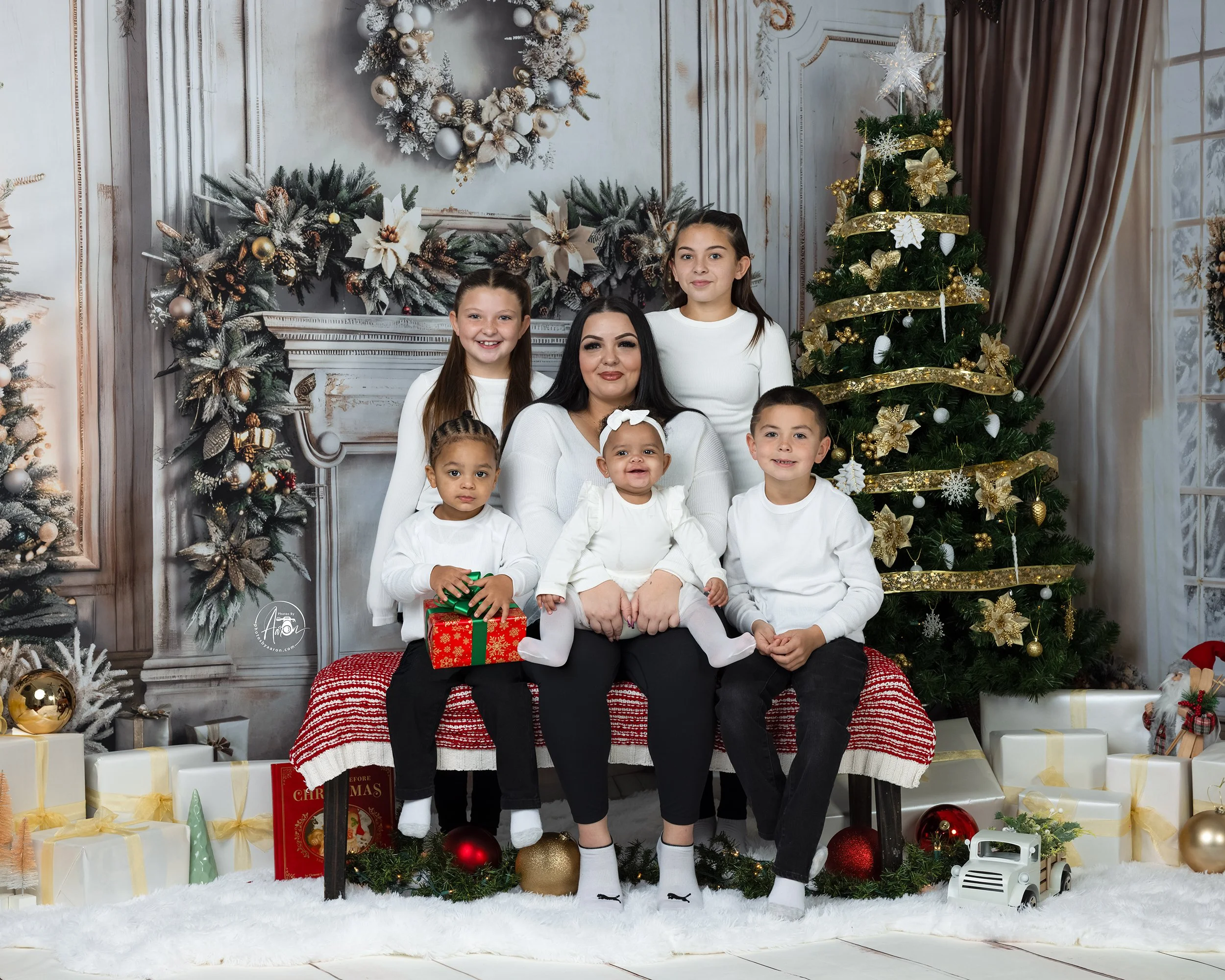 A young boy sitting on a fluffy white rug, opening a golden Christmas gift box. The background includes a fireplace, stockings, and holiday decorations, with a filter of sparkles and glowing hearts around him.