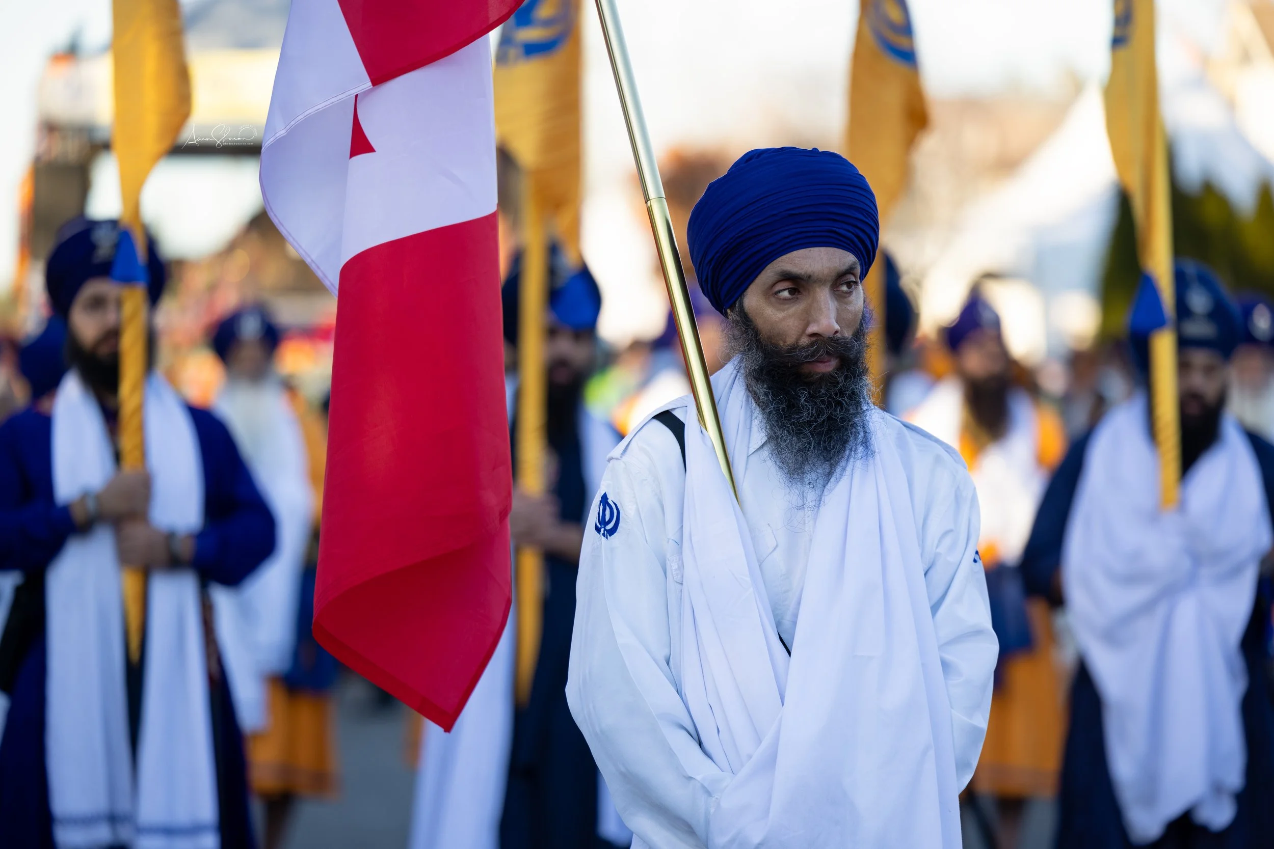 A man wearing a blue turban and white traditional attire holding a Canadian flag in a parade or procession, with other men in similar attire and turbans in the background.