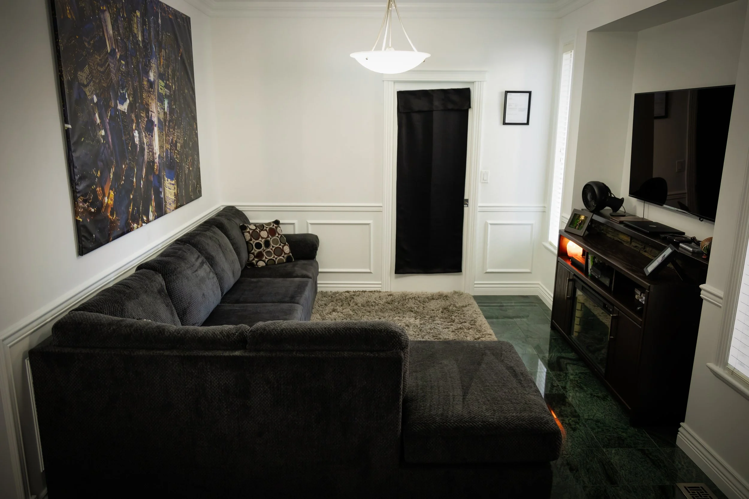 Living room with a dark gray sectional sofa, a television on a black stand, a large cityscape art piece on the wall, a beige rug, a black curtain on the door, and a chandelier overhead.